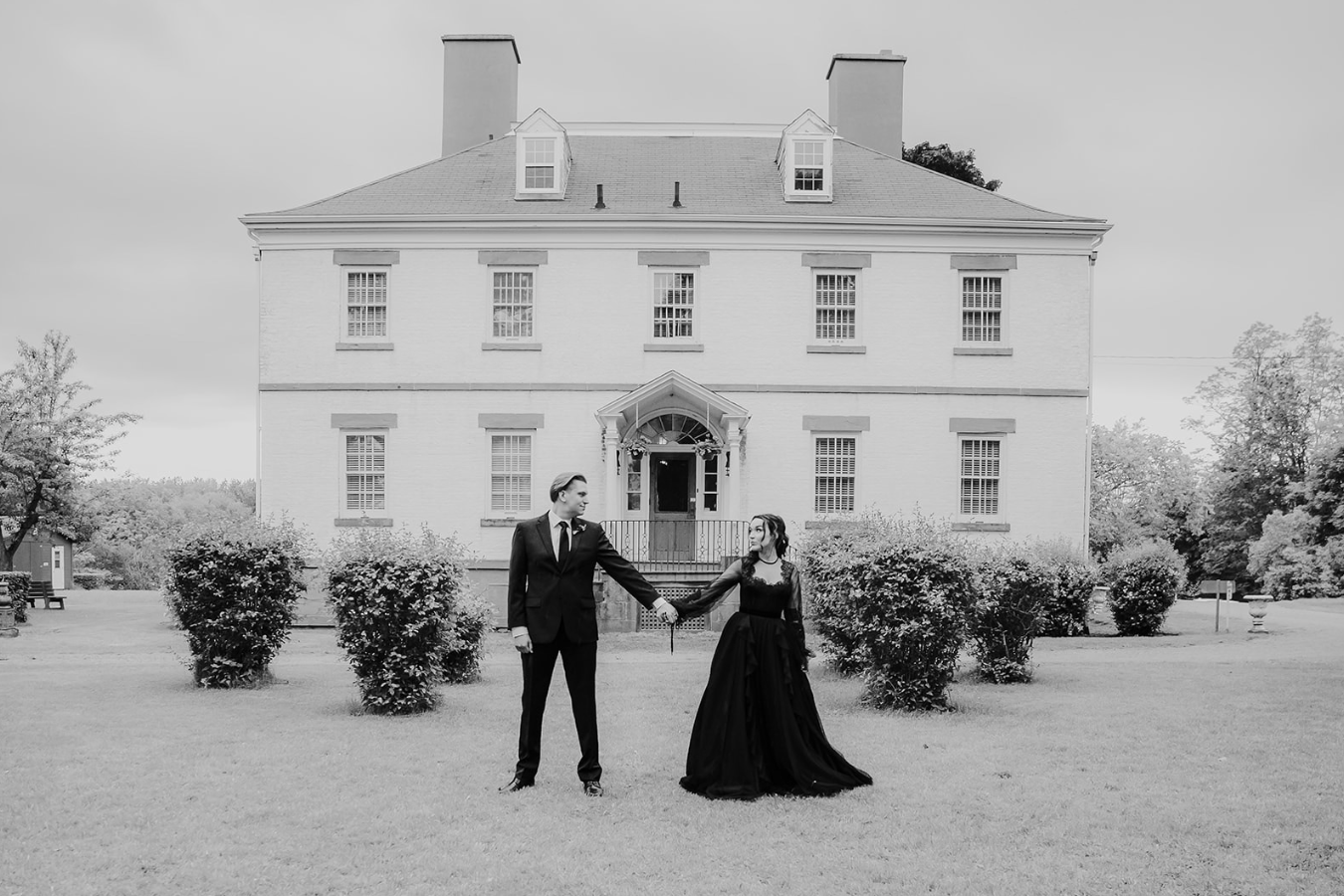 Black and white photo of a couple holding hands in front of a grand white colonial mansion surrounded by manicured shrubs.