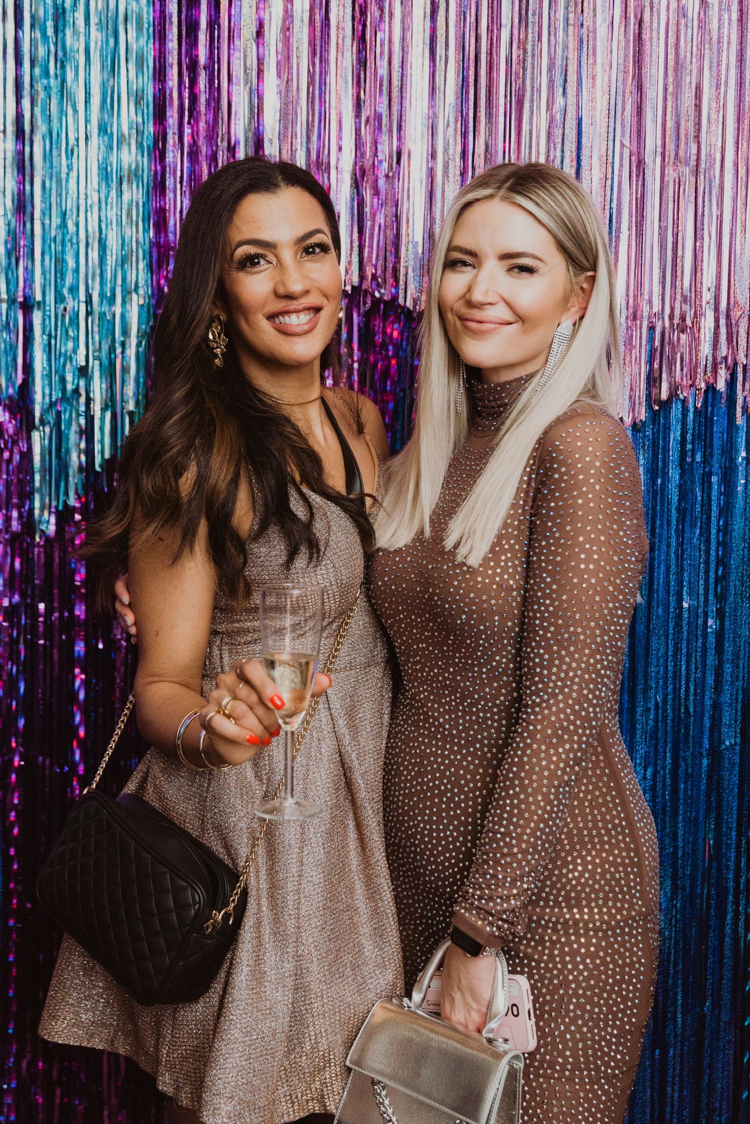 Two women in sparkly dresses posing together at a party in front of a colorful tinsel backdrop.