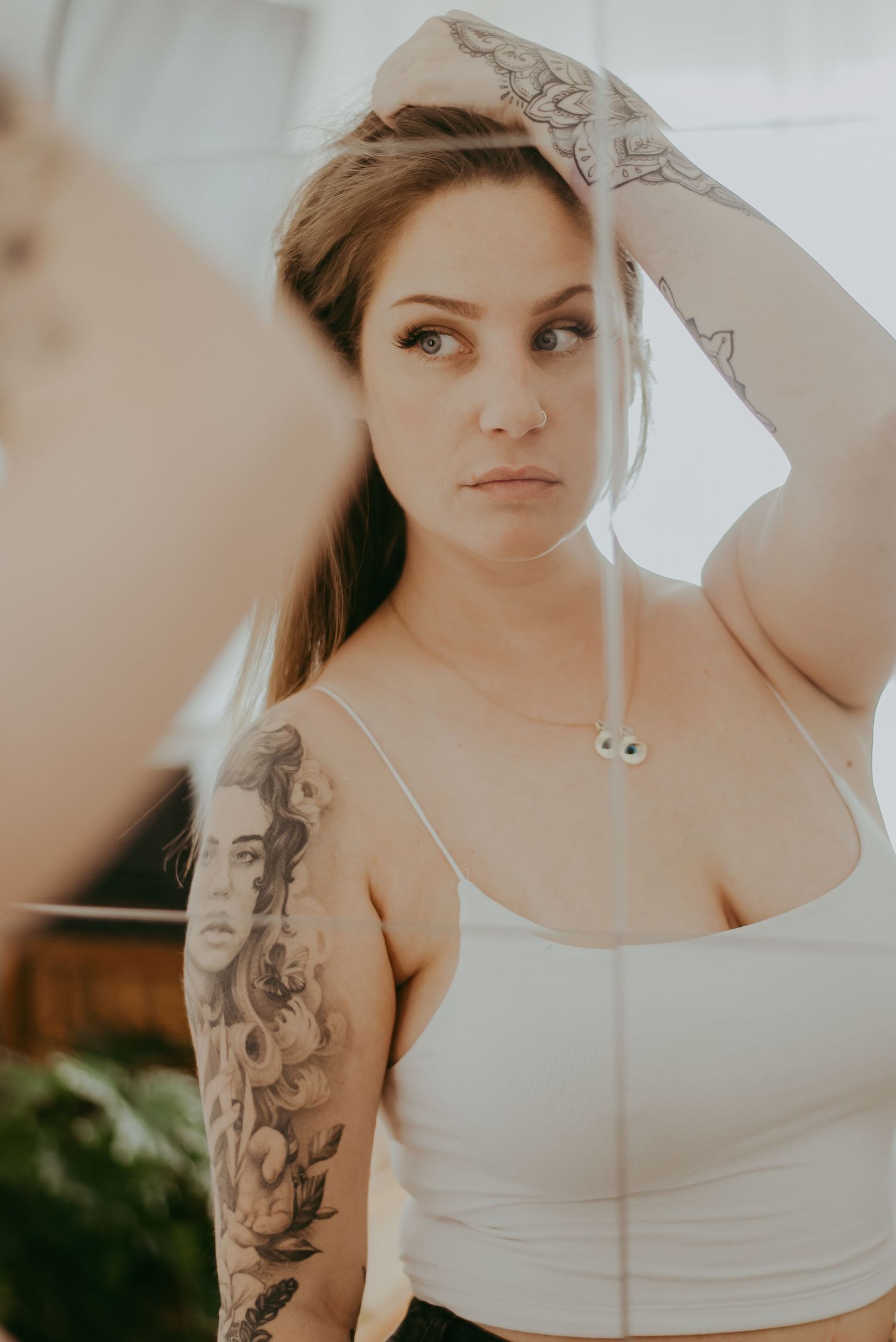 Woman with sleeve tattoos in white crop top poses confidently, hand raised to head, in soft natural light.