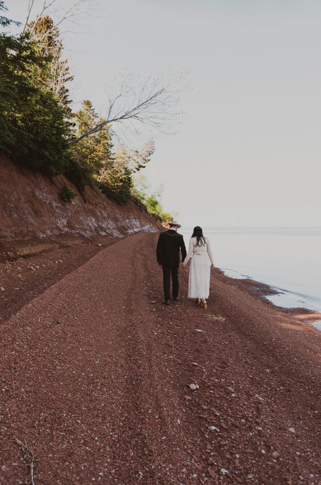 A couple in wedding attire walks hand-in-hand along a scenic red gravel path beside calm water.