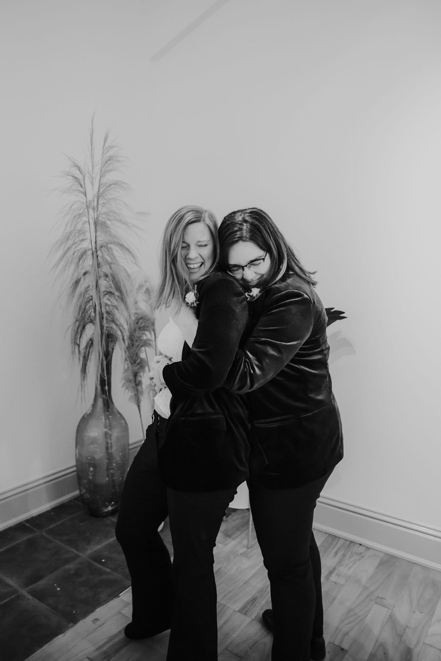 Two women hugging and laughing in black and white photo with decorative plant in background.