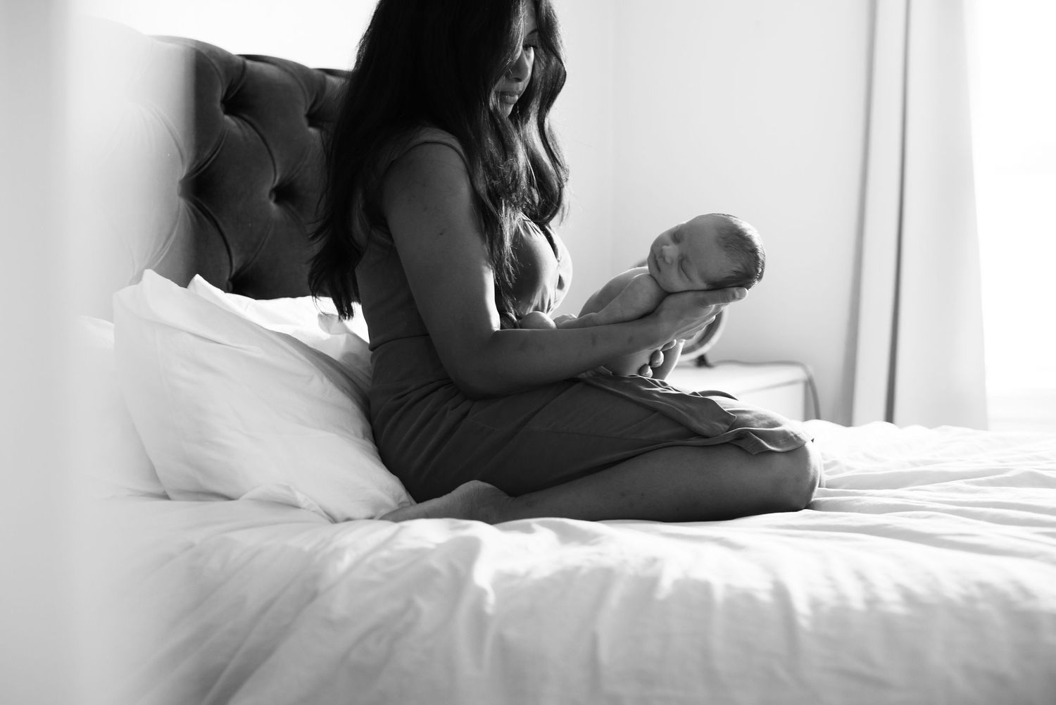 Black and white photo of a mother sitting on a bed cradling her newborn baby in a serene bedroom.