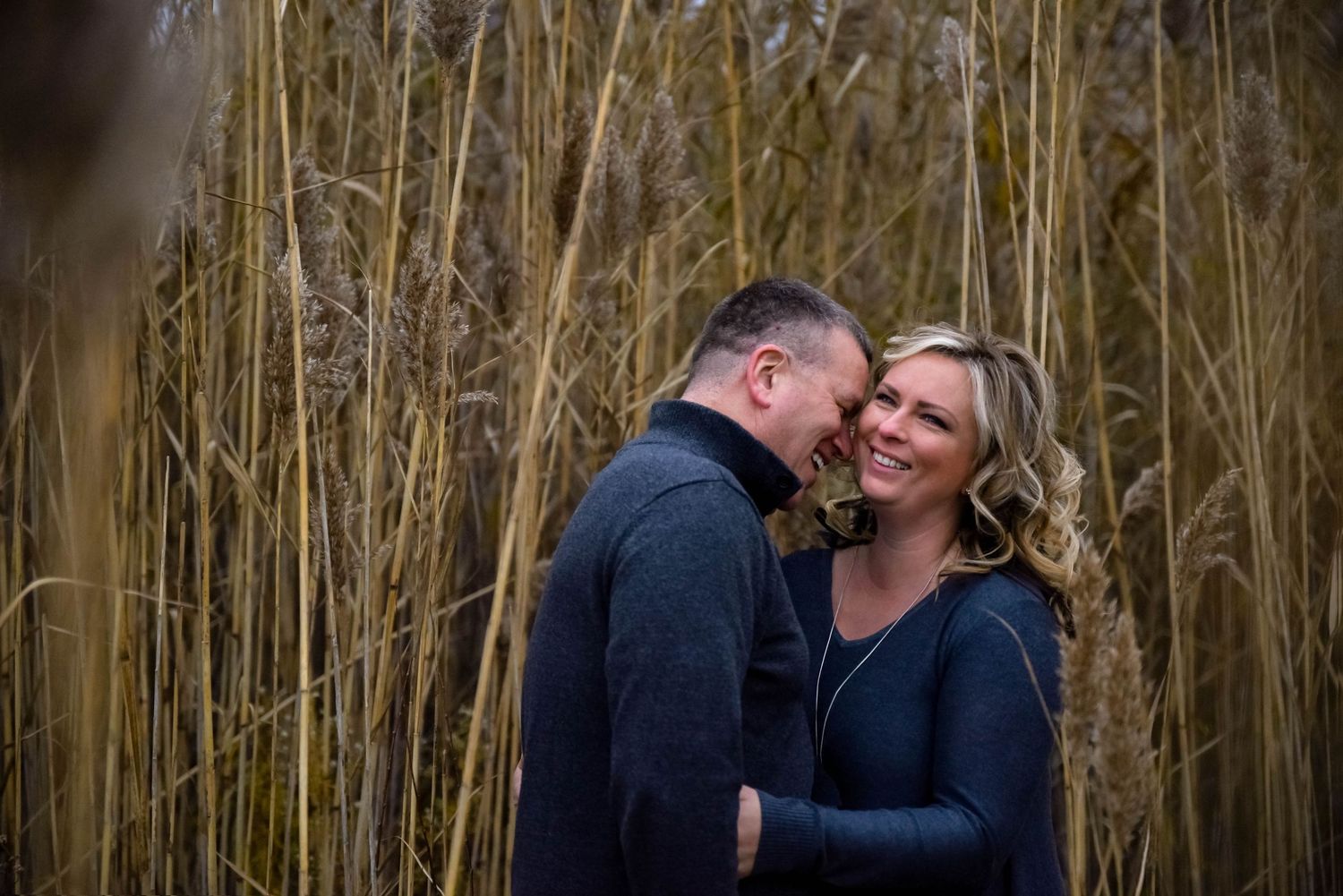 Couple embracing and smiling among tall golden reeds in an outdoor engagement photo session.