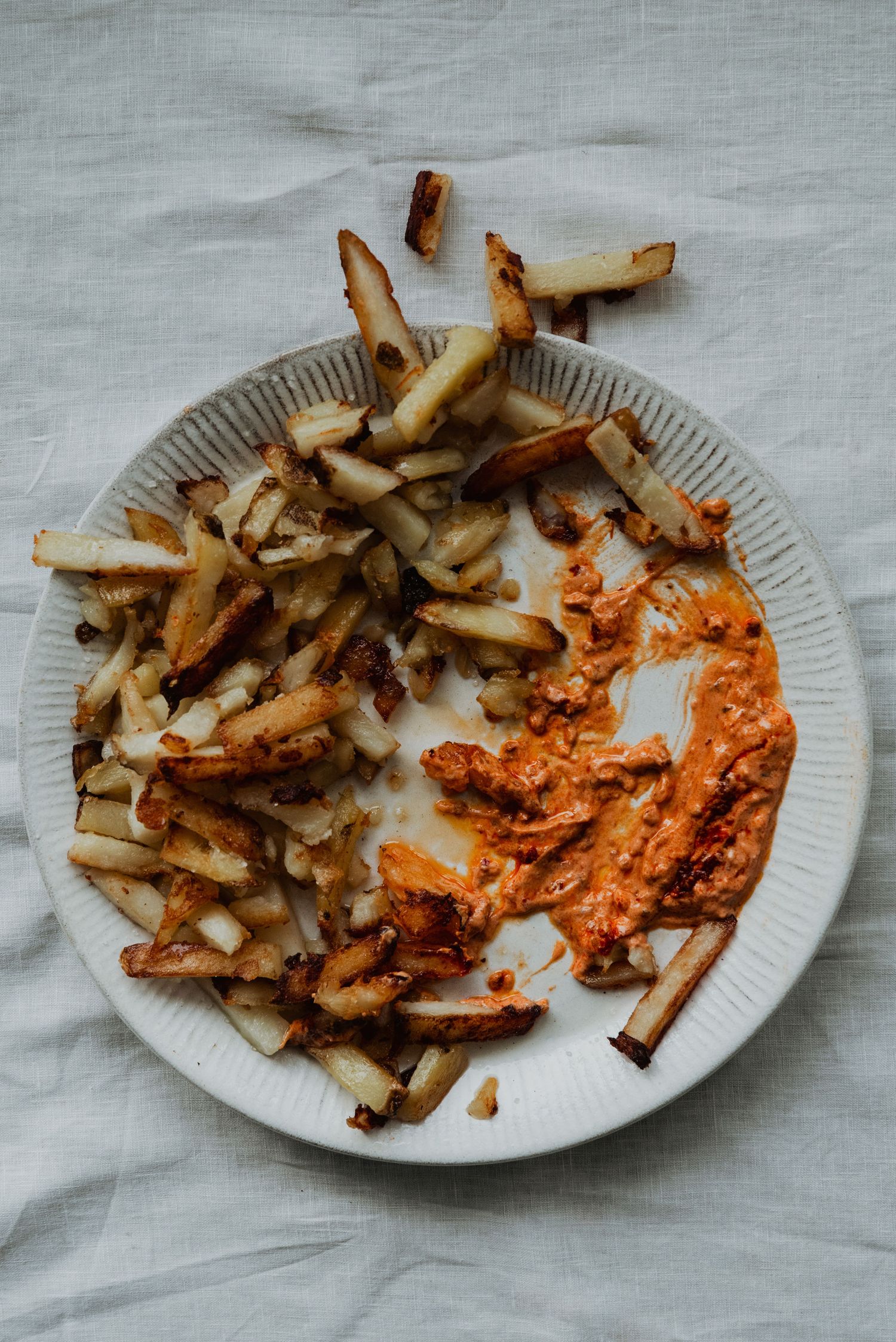 A white ceramic plate with homemade french fries and a side of spicy orange sauce on a linen cloth.