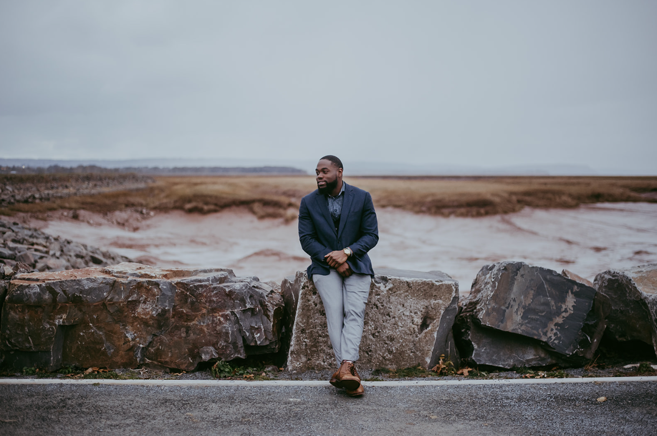 Man in navy blazer and gray pants leaning on rocks by a coastal shoreline on an overcast day.