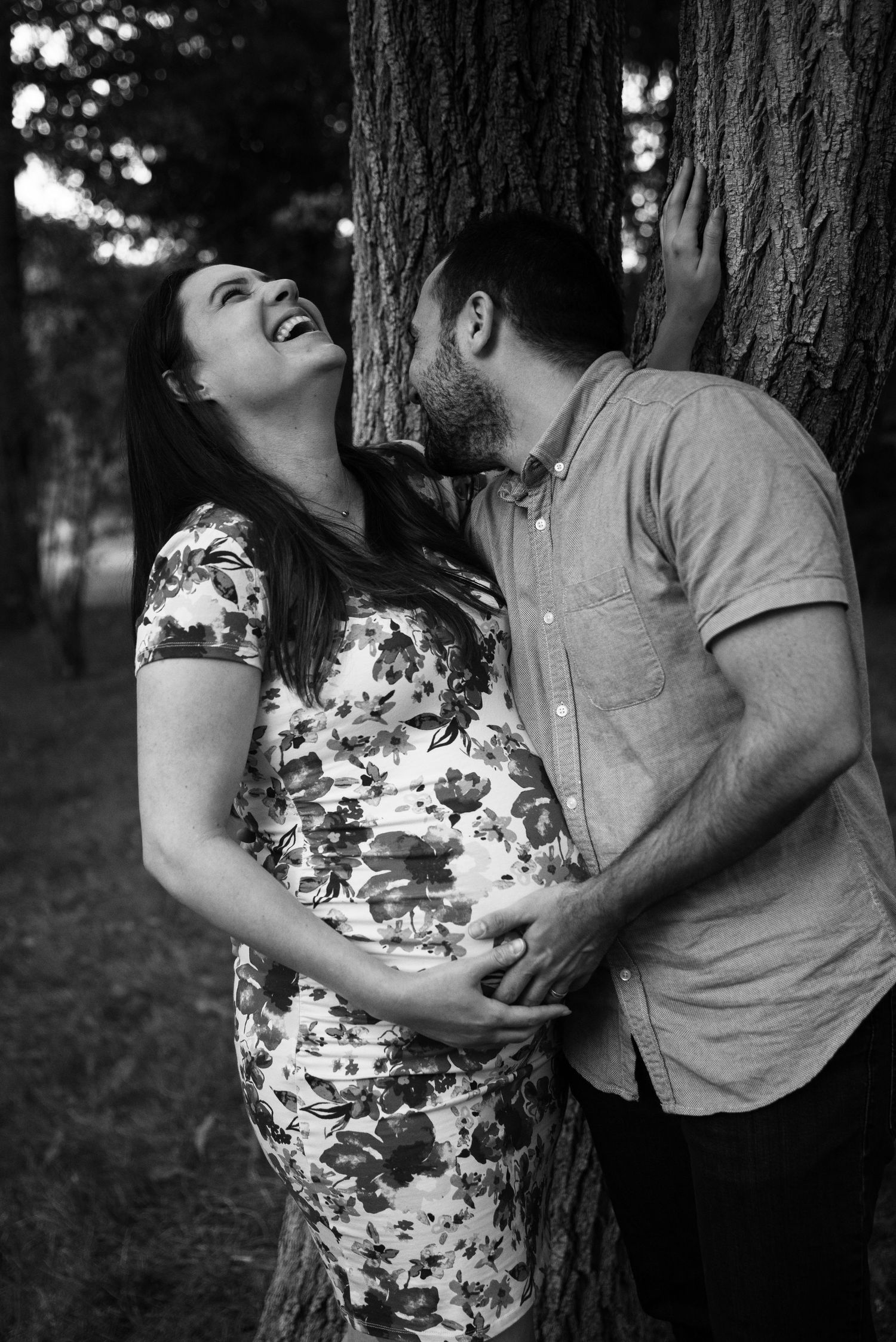 Happy pregnant couple laughing and embracing against a tree in a black and white maternity photo.