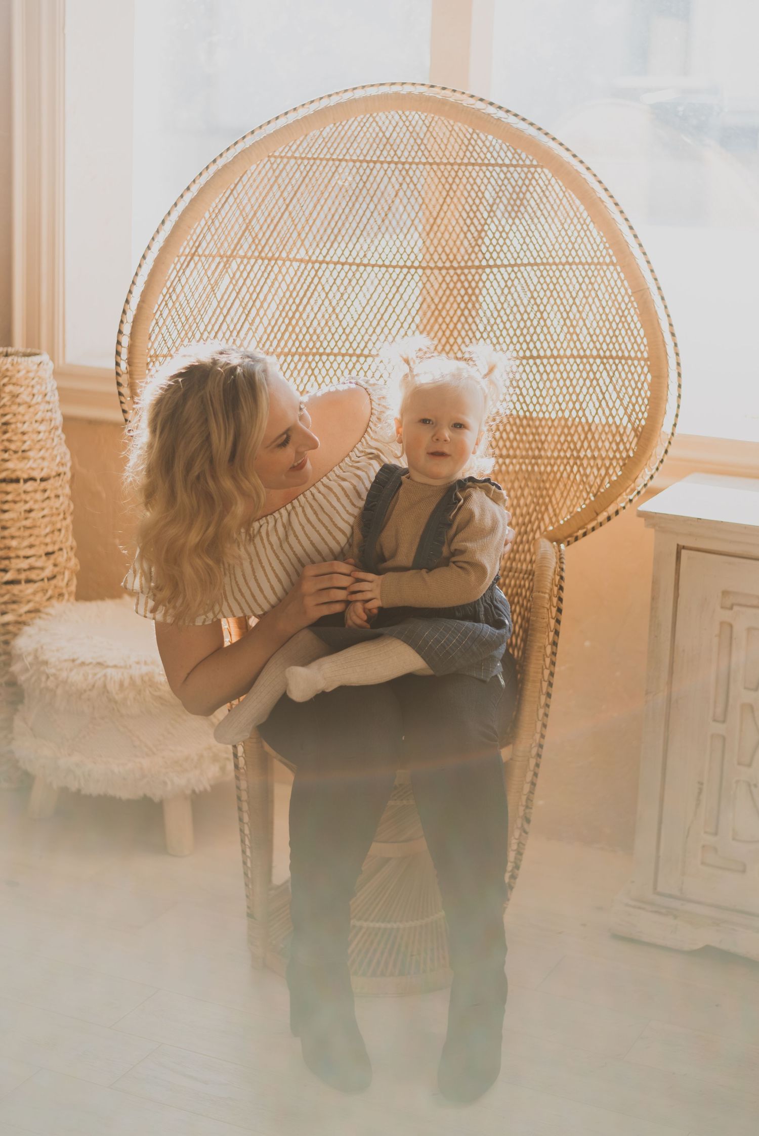 A smiling mother holds her baby girl in a large wicker peacock chair by a bright window.