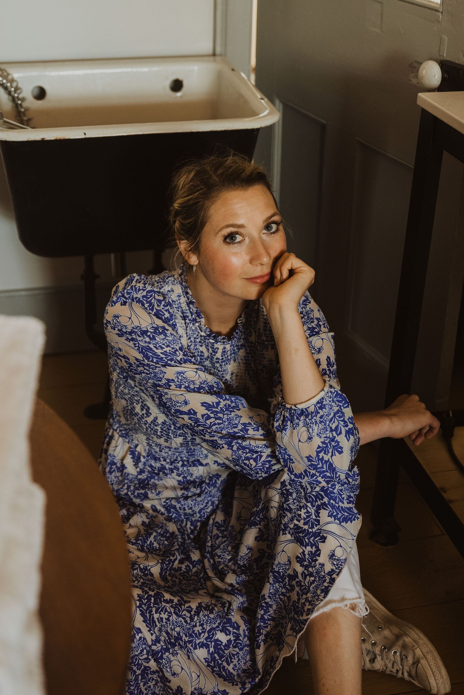 Woman in blue floral robe sitting on floor leaning against bathroom sink, looking up thoughtfully.