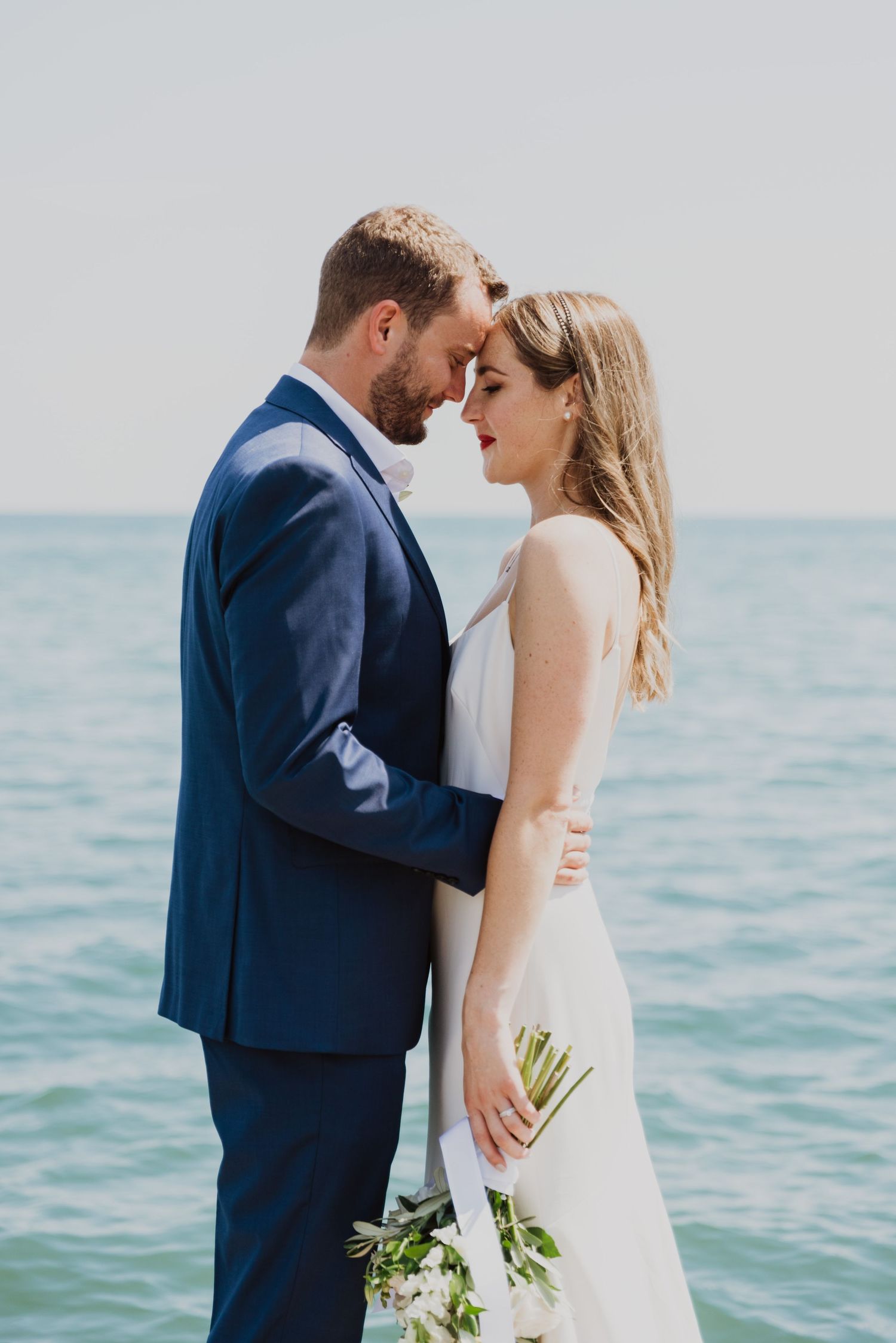 Bride and groom share an intimate moment by the ocean, she holds a bouquet in a white gown as he wears a navy suit.