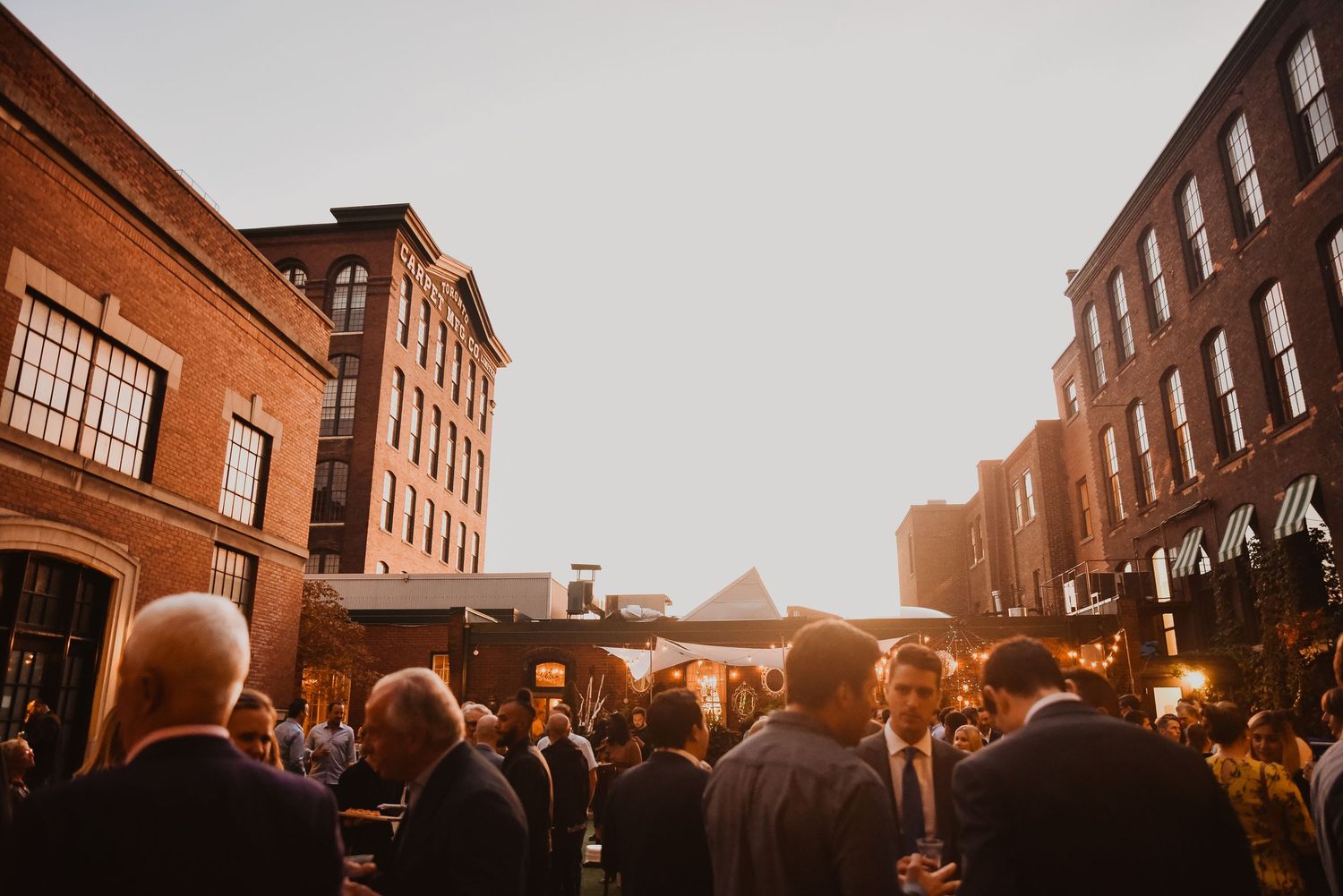 Guests mingle outdoors at golden hour between historic brick buildings at an elegant evening event.