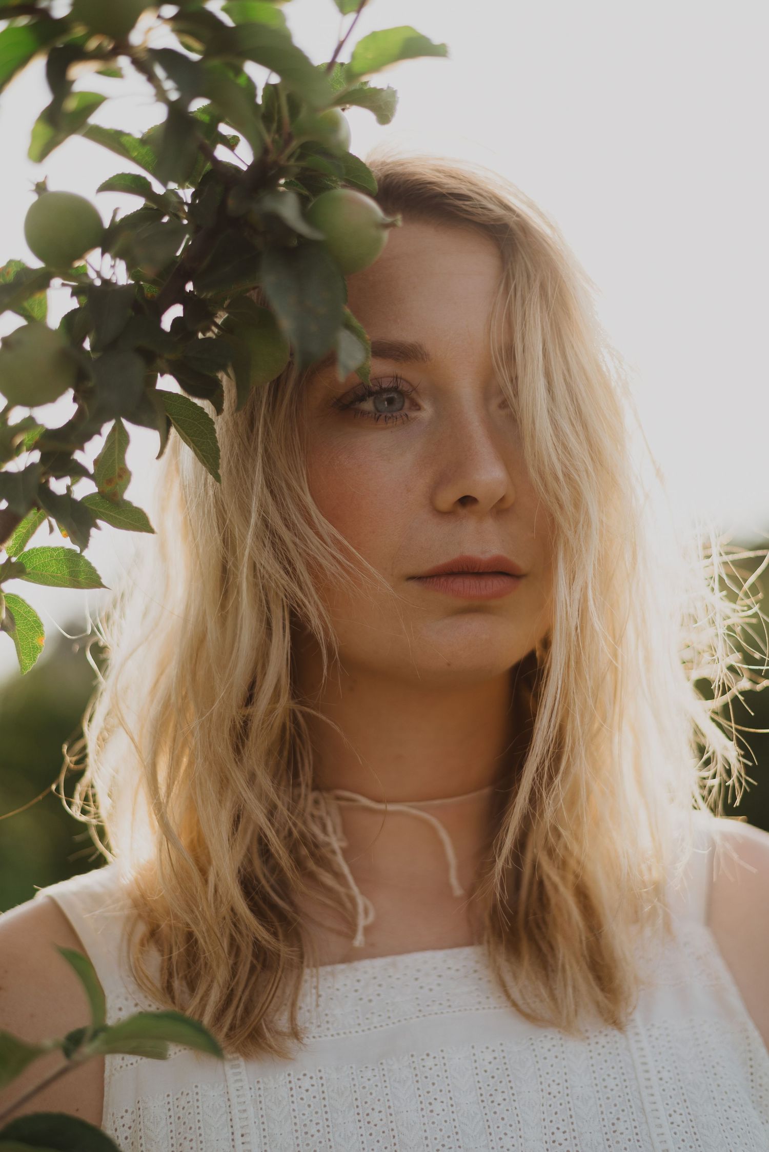 Young blonde woman in white top gazes softly beside green apple tree branches in natural outdoor light.