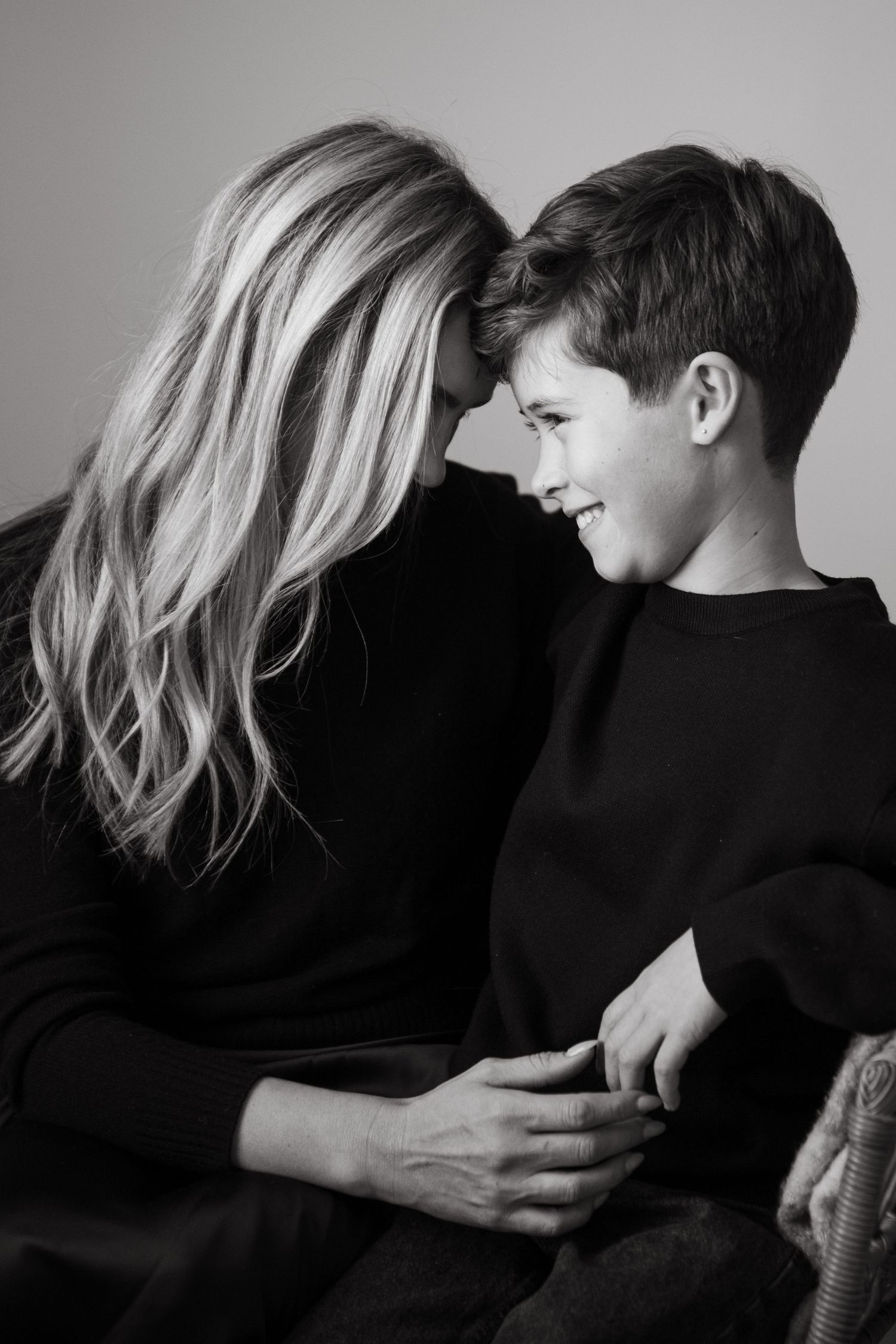 Black and white photo of a smiling boy and blonde woman in black sweaters sharing a tender forehead-to-forehead moment.