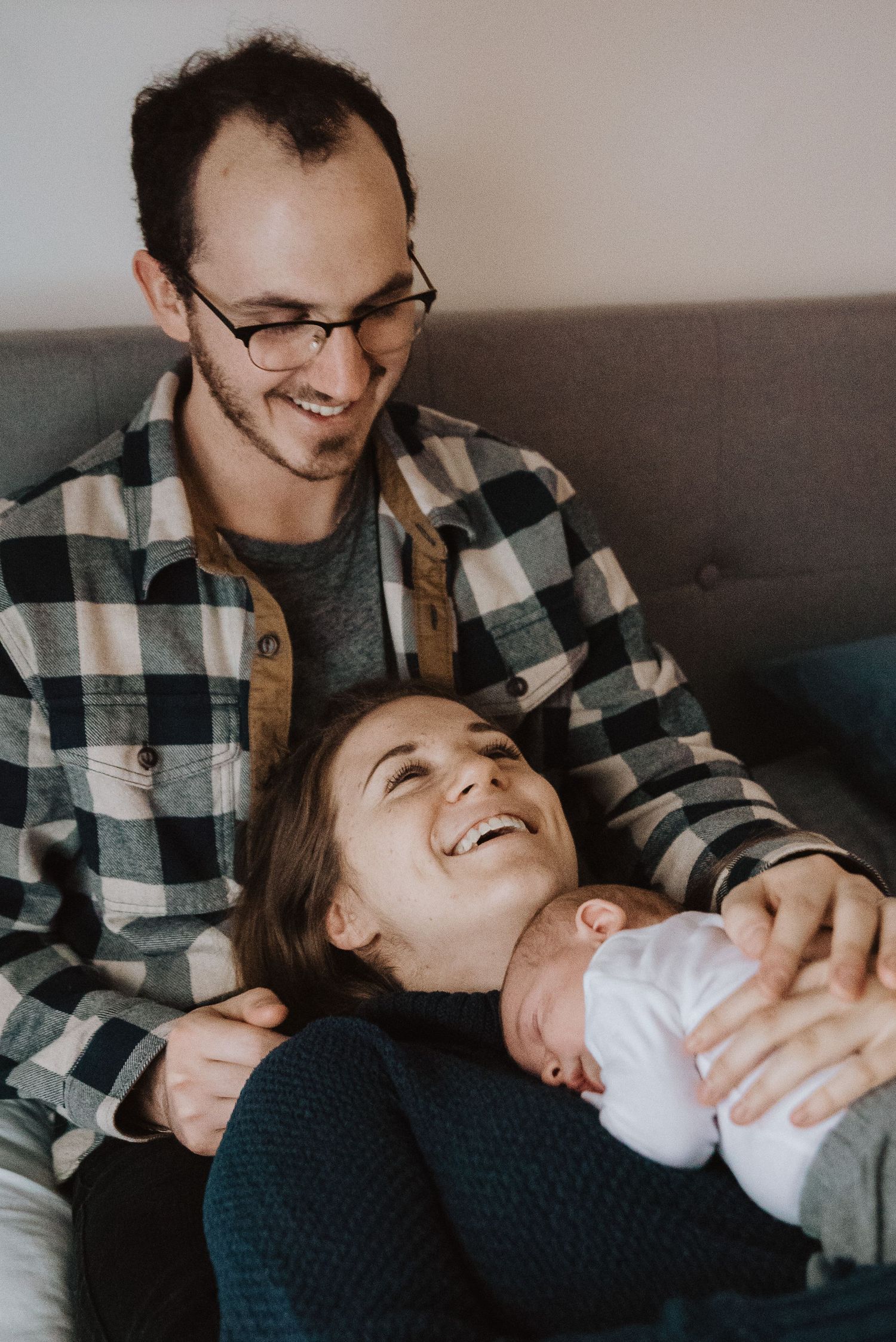 Happy young parents smiling and holding their newborn baby together on a cozy couch at home.
