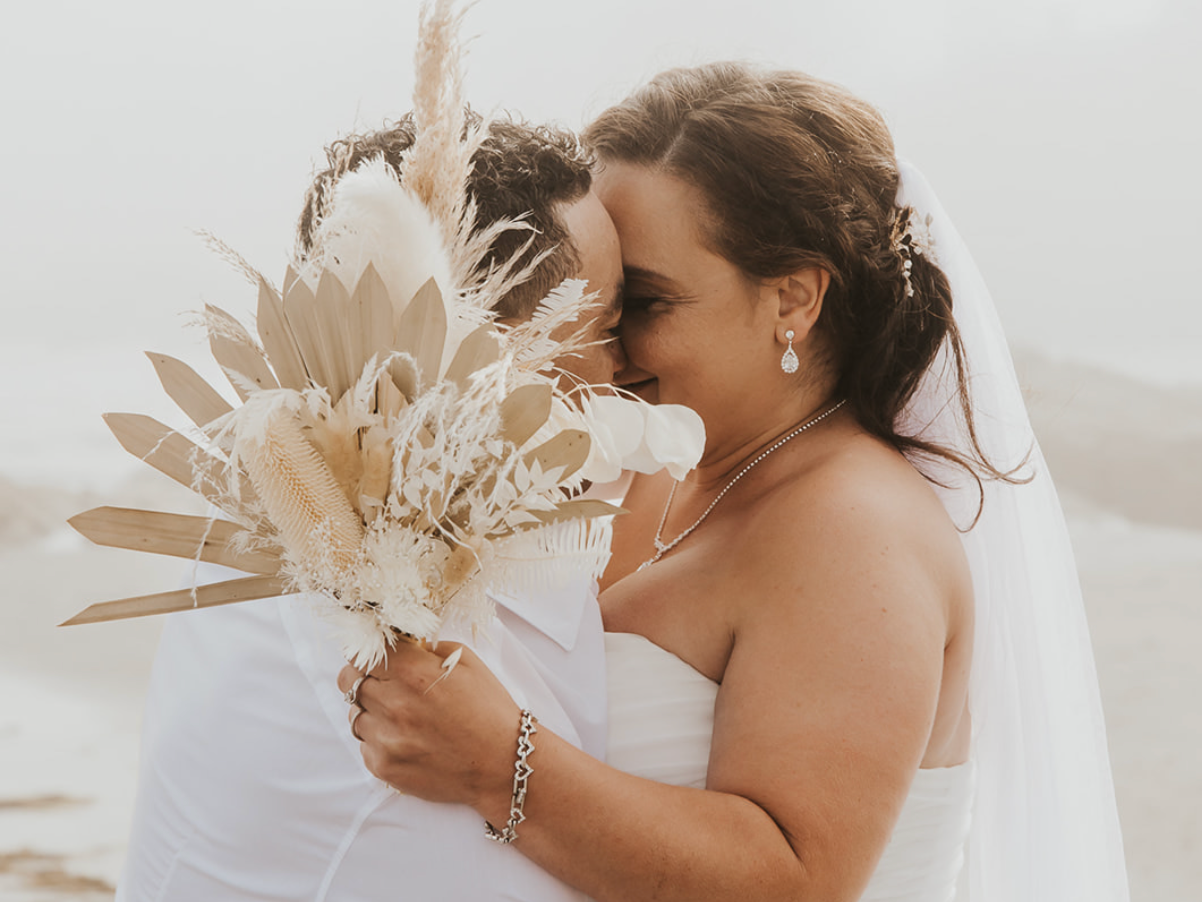 Bride and groom sharing an intimate moment, bride holding a boho dried floral bouquet in neutral tones at a beach wedding.