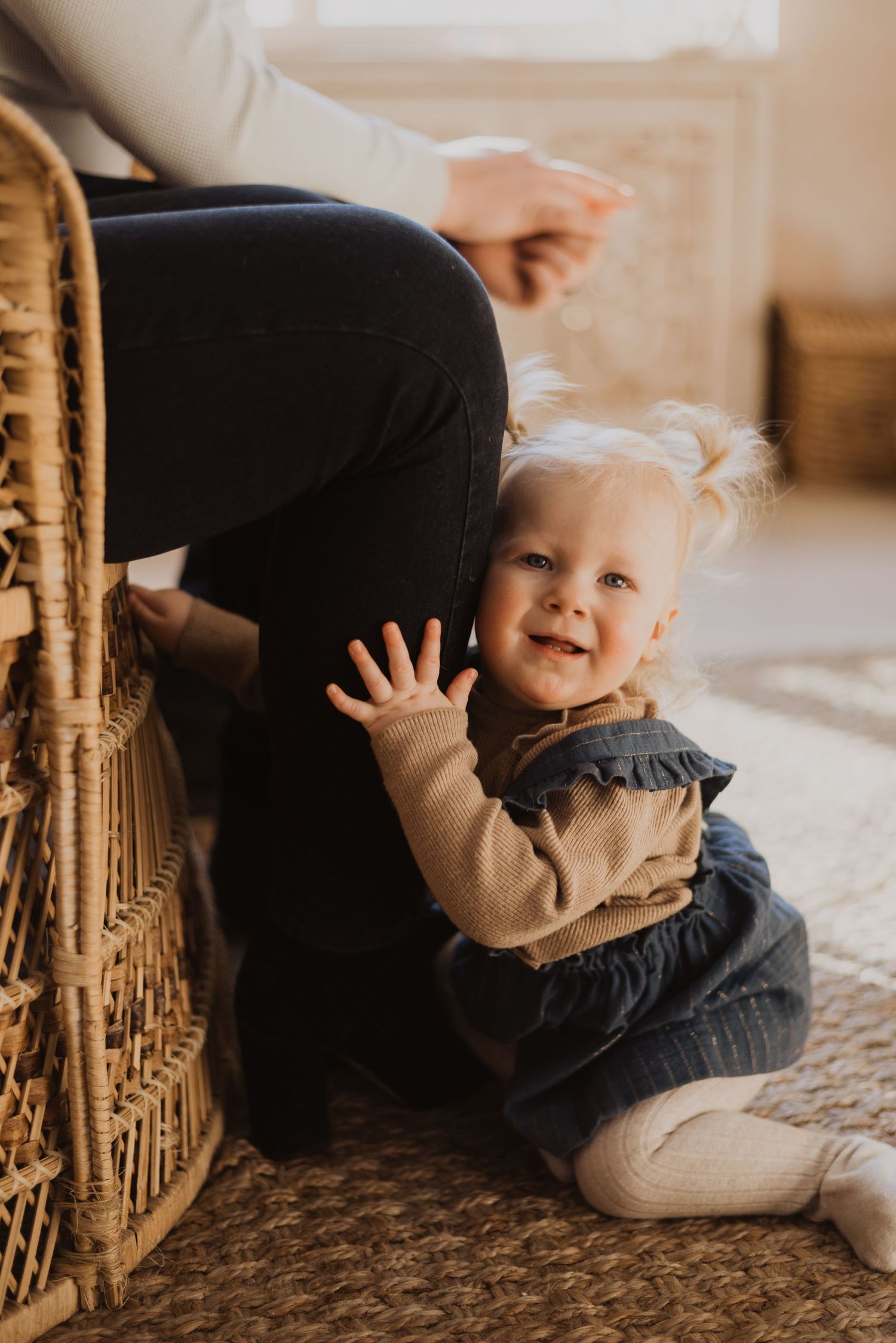 Smiling baby girl in denim dress sitting on floor, waving hand beside wicker chair with adult nearby.