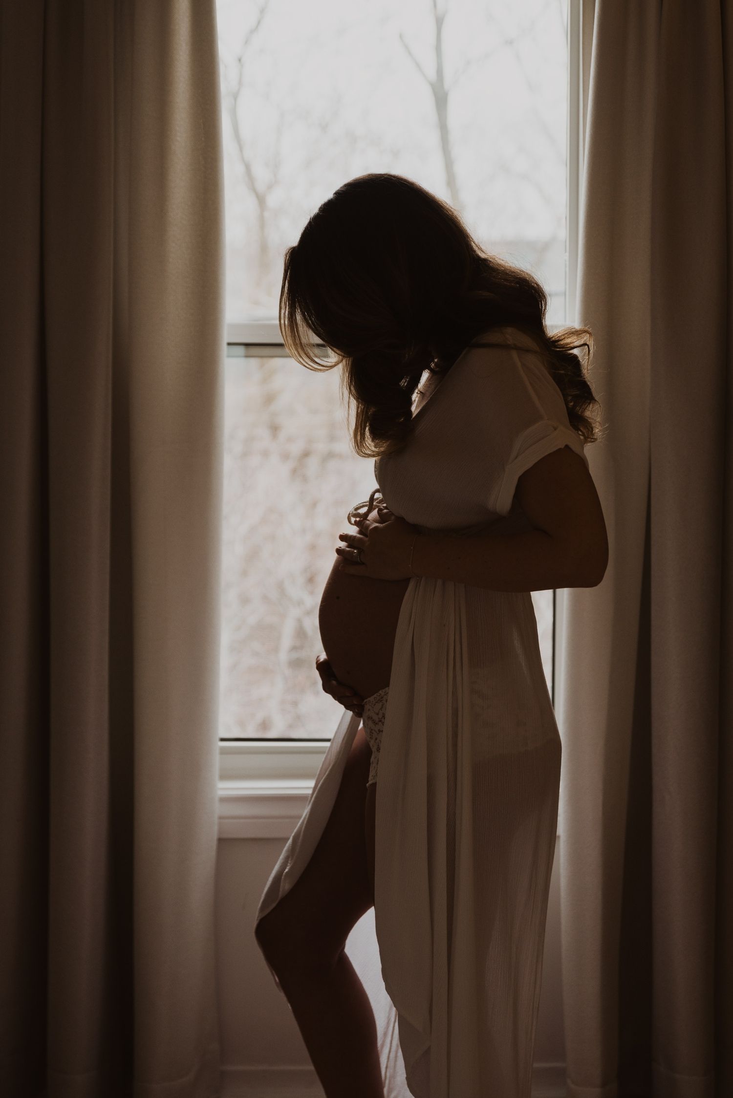 Pregnant woman in white dress cradling baby bump by window with soft backlight and curtains.
