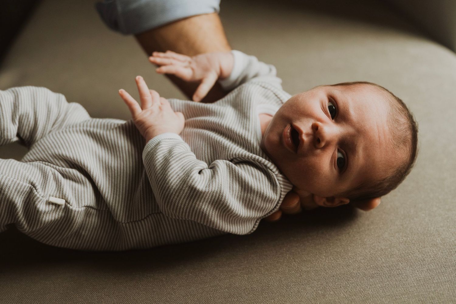 Newborn baby in striped onesie lying on back, looking up with bright eyes while adult hand gently supports them.