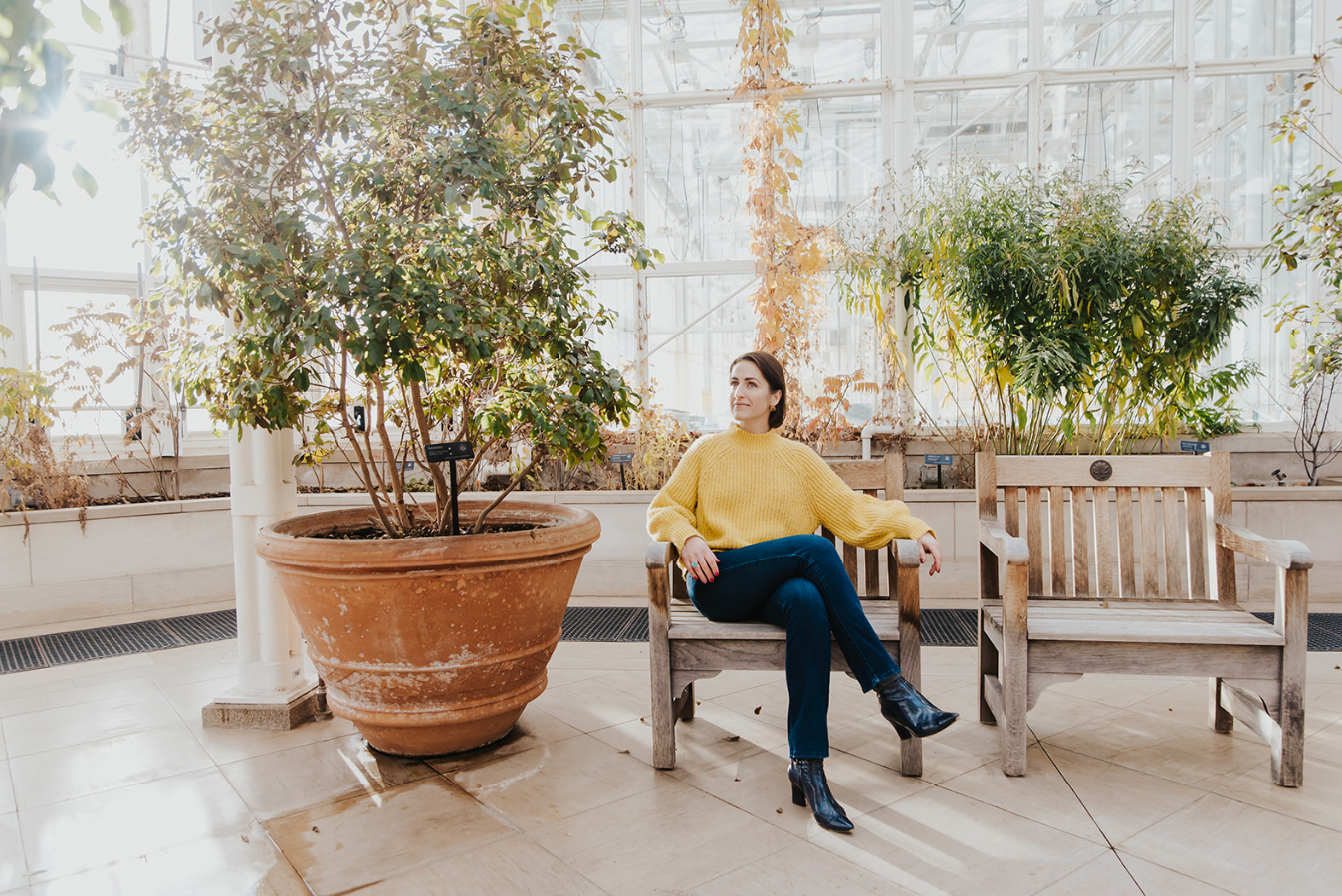 Woman in yellow sweater sitting on wooden bench surrounded by lush green plants in a bright greenhouse.