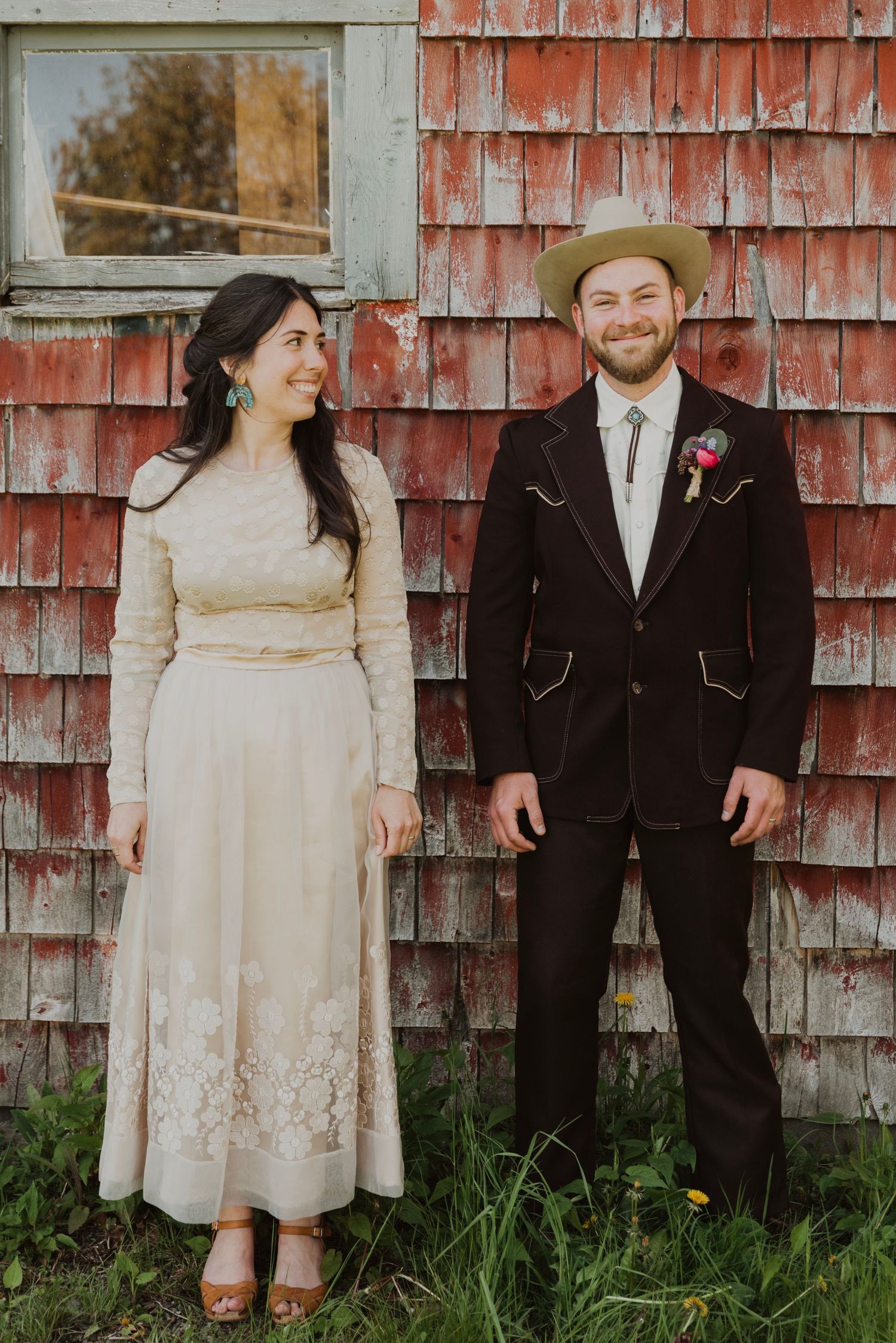 A couple in vintage wedding attire stands smiling before a rustic red barn wall surrounded by green grass.