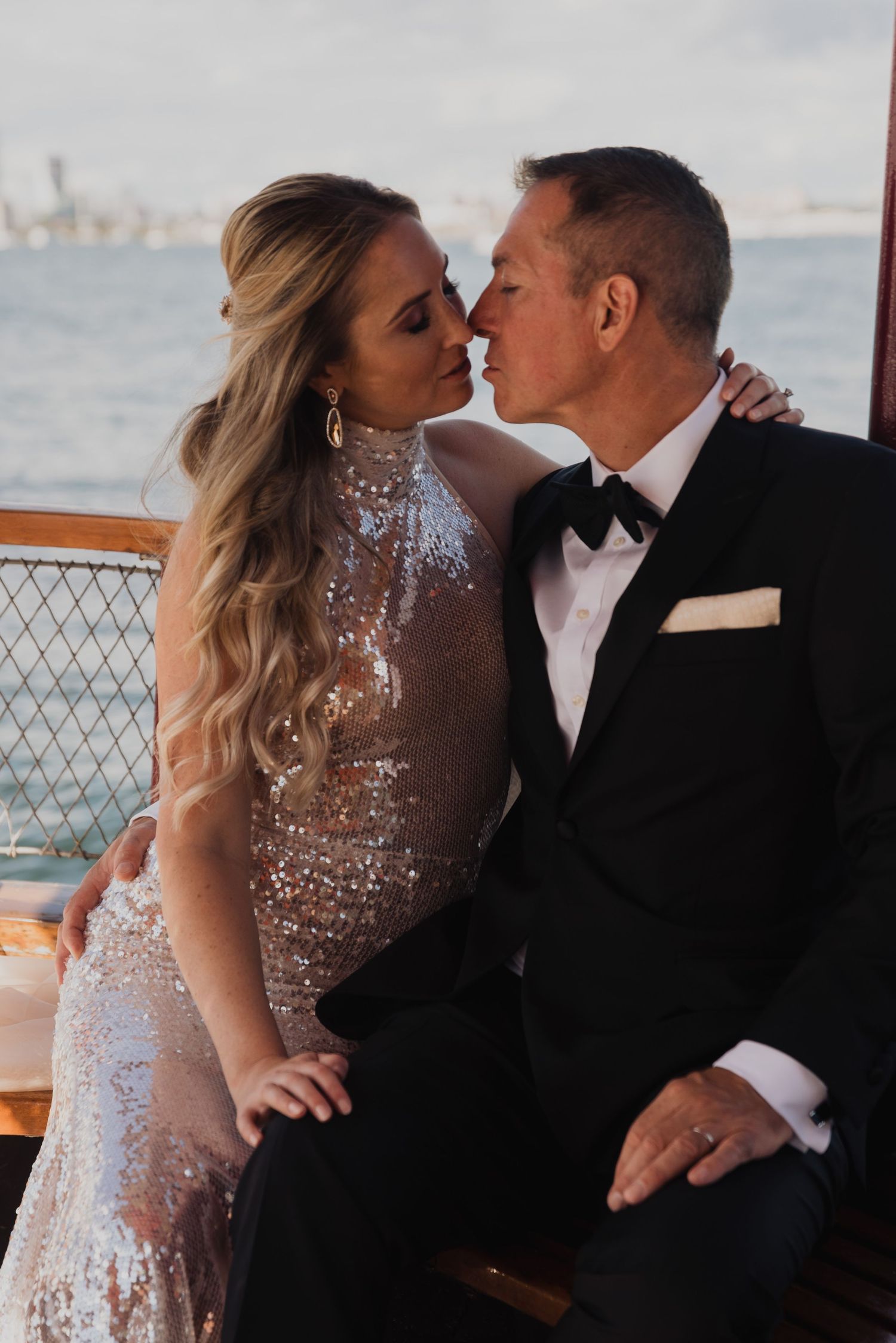 Bride in sequin gown and groom in tuxedo share romantic moment on a boat with ocean backdrop.