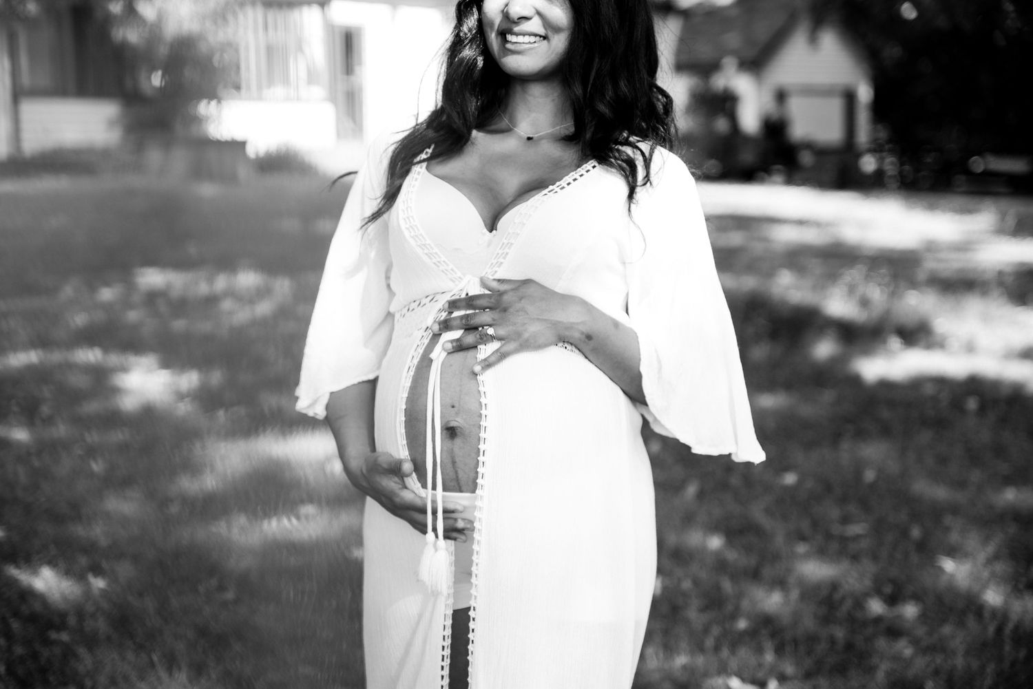 Black and white maternity photo of a smiling woman in a white dress cradling her baby bump outdoors.