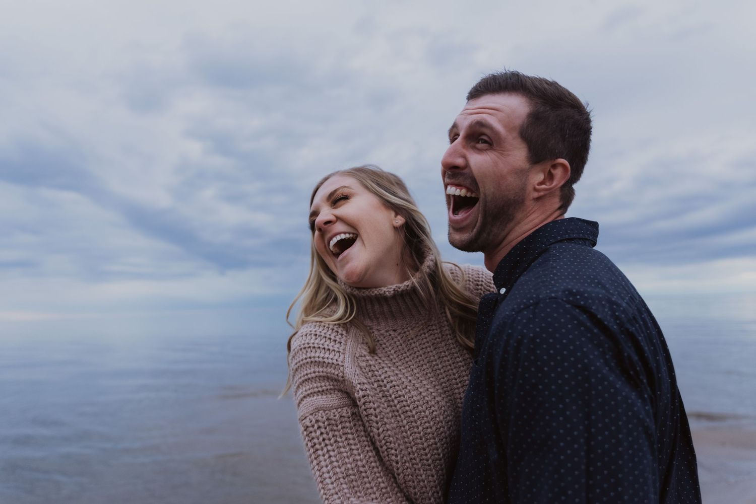 A joyful couple laughing together outdoors near the ocean under a cloudy sky, wearing cozy sweaters.