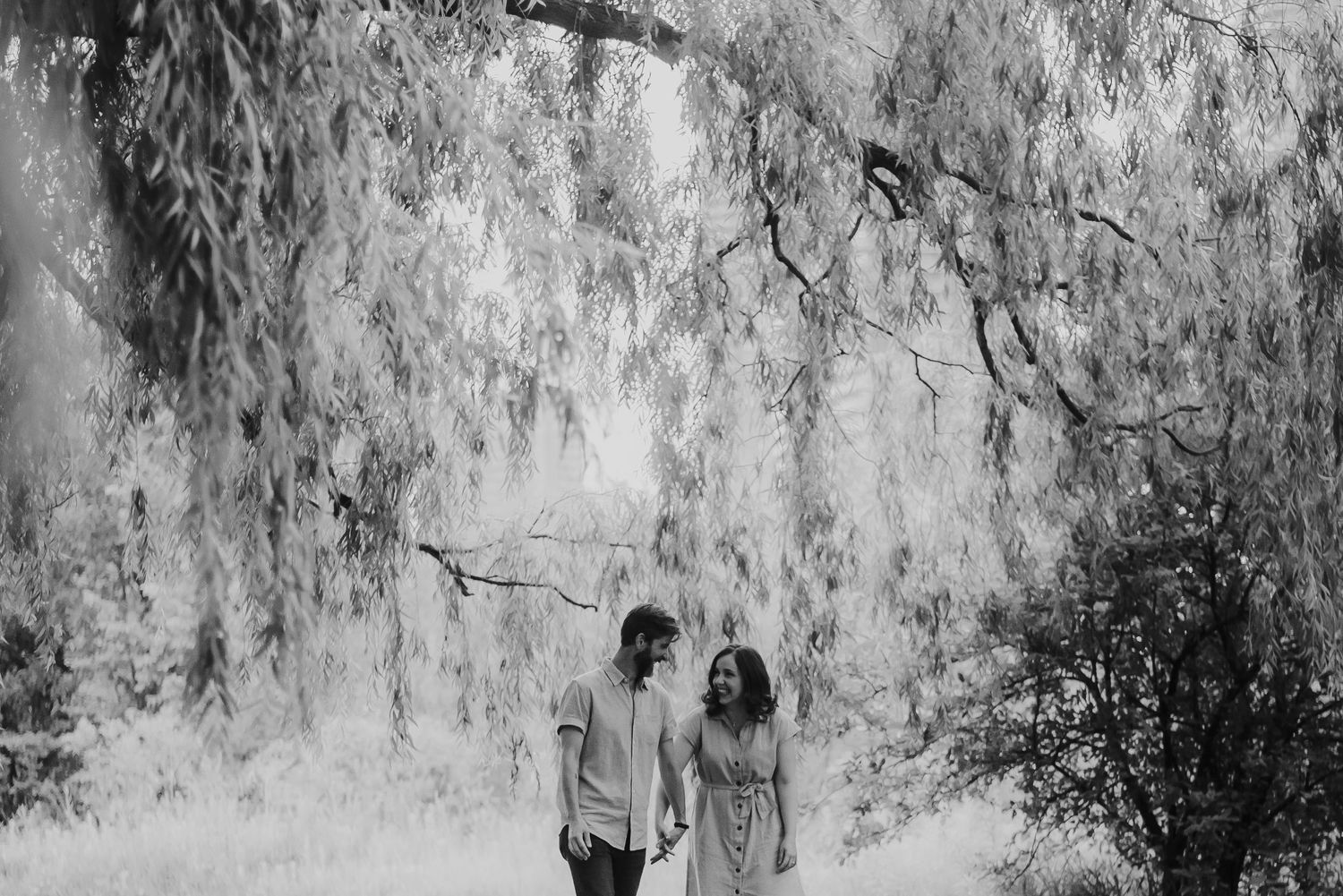 Black and white photo of a couple walking hand in hand beneath weeping willow branches in a serene forest setting.