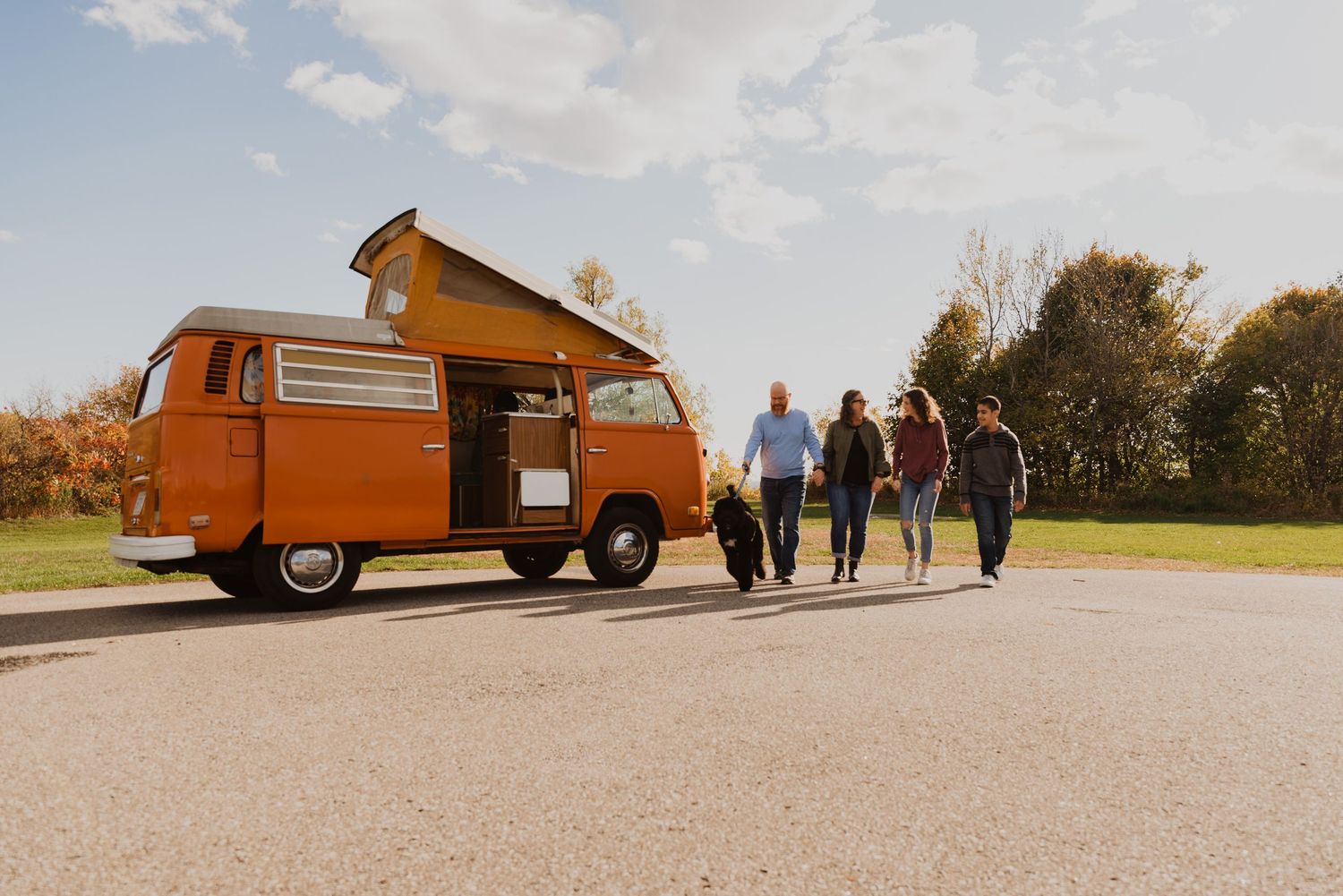 Group of friends walking toward a vintage orange VW camper van on a sunny day in an open parking area.