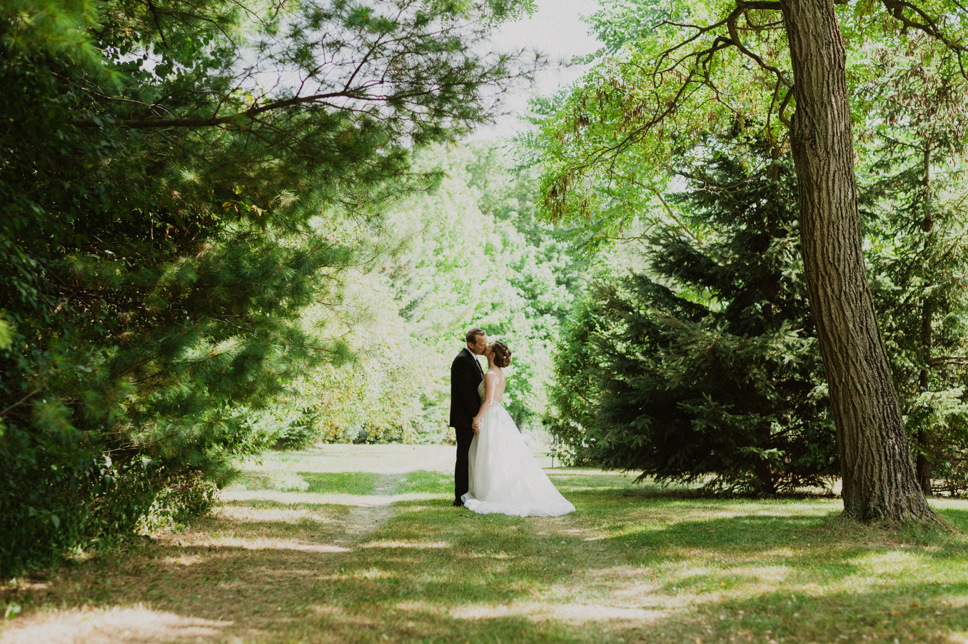 A bride and groom share a romantic kiss surrounded by lush green trees in a sunlit outdoor garden setting.