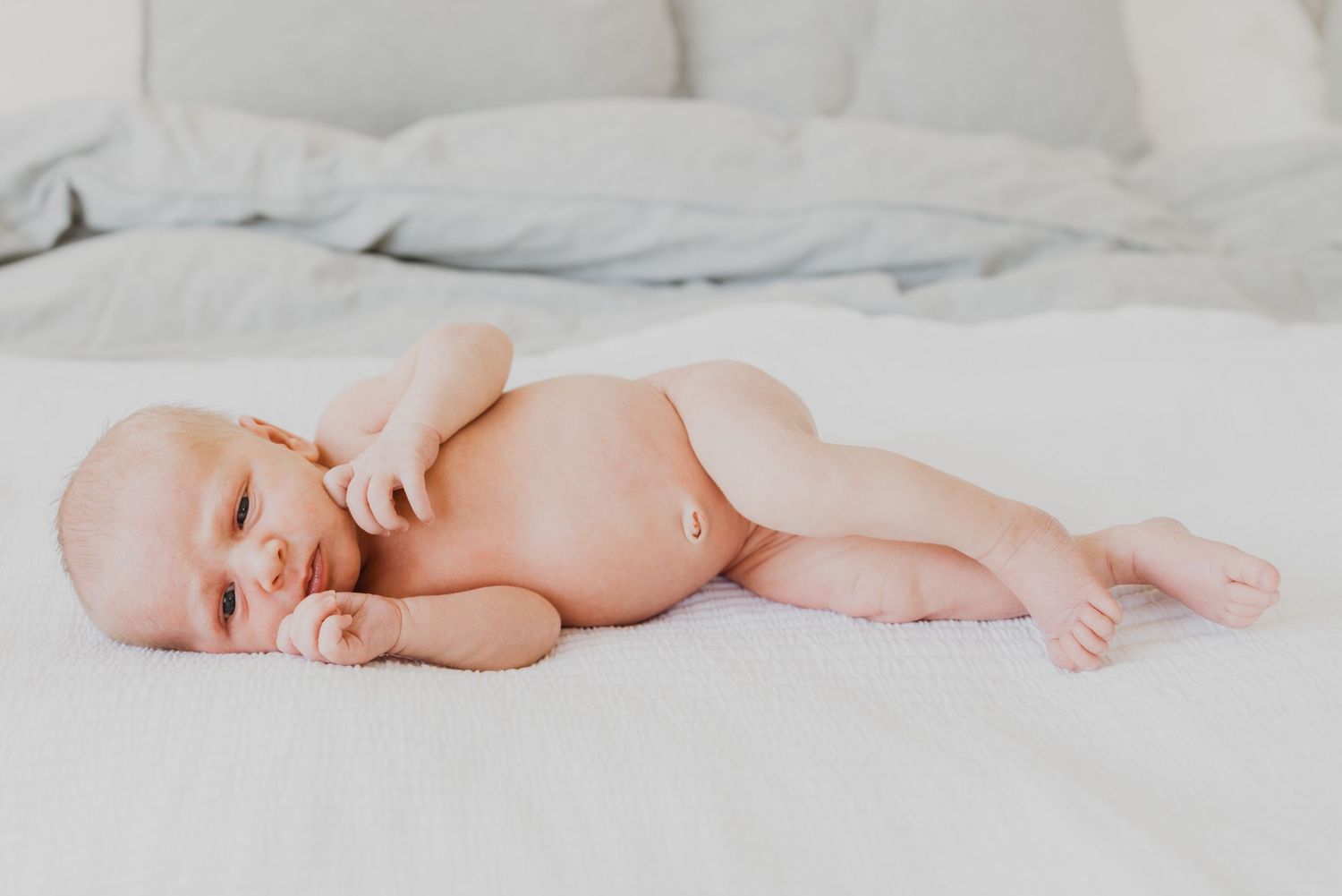 Newborn baby lying on a white bed in a diaper, peacefully resting with eyes closed in a soft, bright setting.