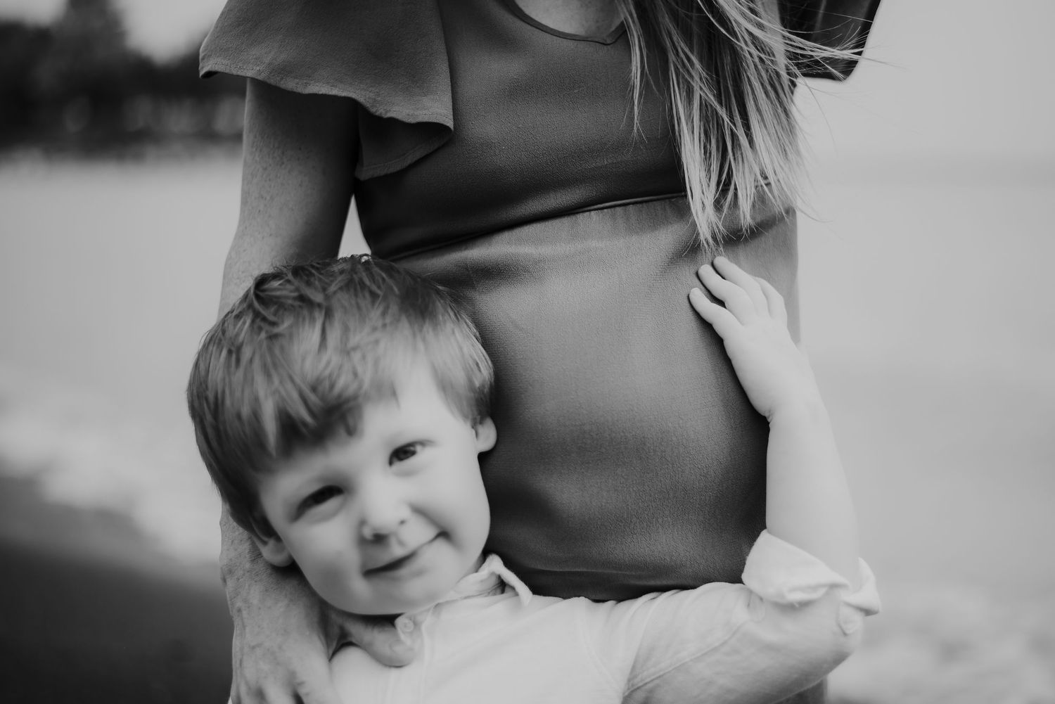 Black and white photo of a smiling toddler leaning against a pregnant woman's belly outdoors.