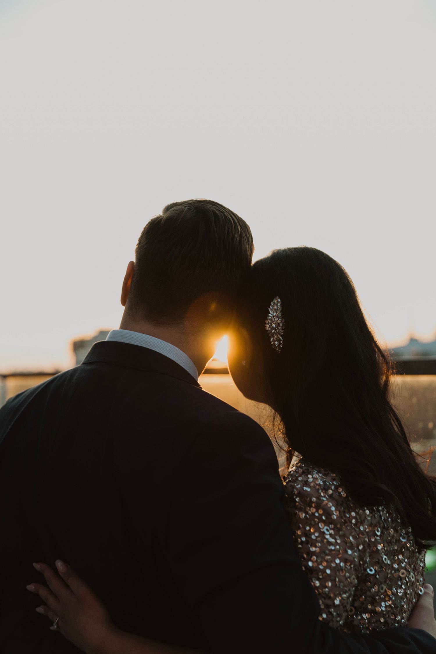 Romantic couple silhouetted against golden sunset light, woman wearing sparkly dress and jeweled hair accessory.
