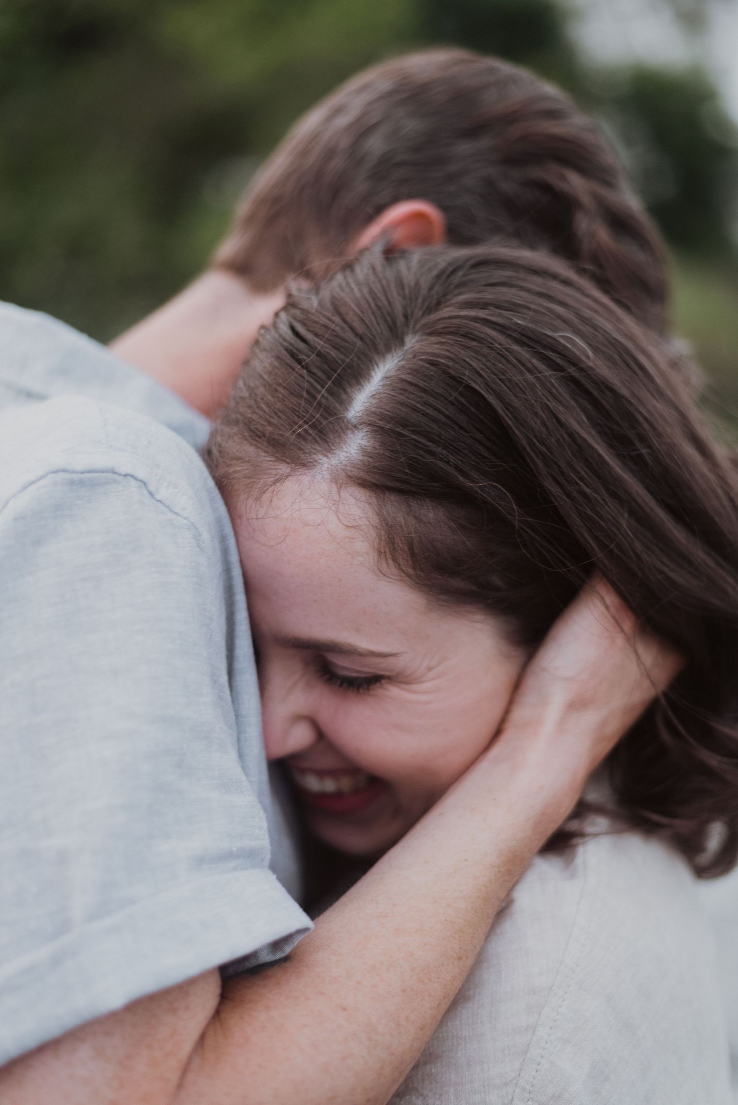A smiling woman hugs a man in a light gray t-shirt outdoors, resting her head on his shoulder in a tender embrace.