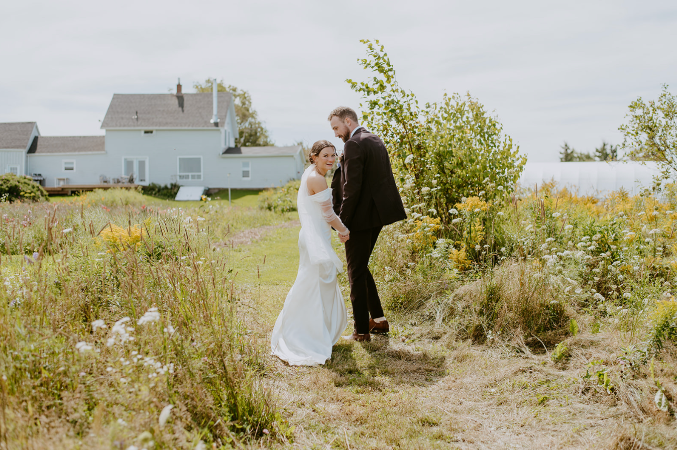 Bride and groom stand on a wildflower meadow path near a white house and waterfront on their wedding day.