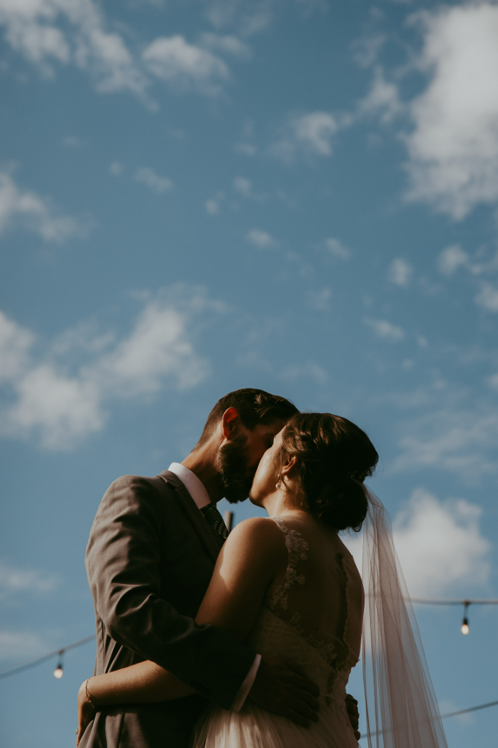 A bride and groom share a romantic kiss under a blue sky with white clouds at their outdoor wedding ceremony.