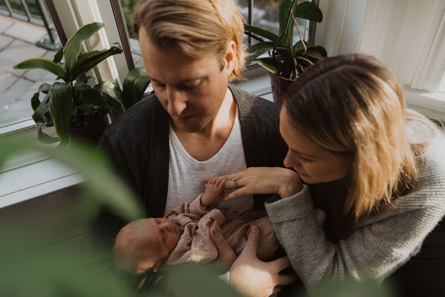 New parents lovingly cradling their newborn baby by a sunlit window surrounded by green plants.