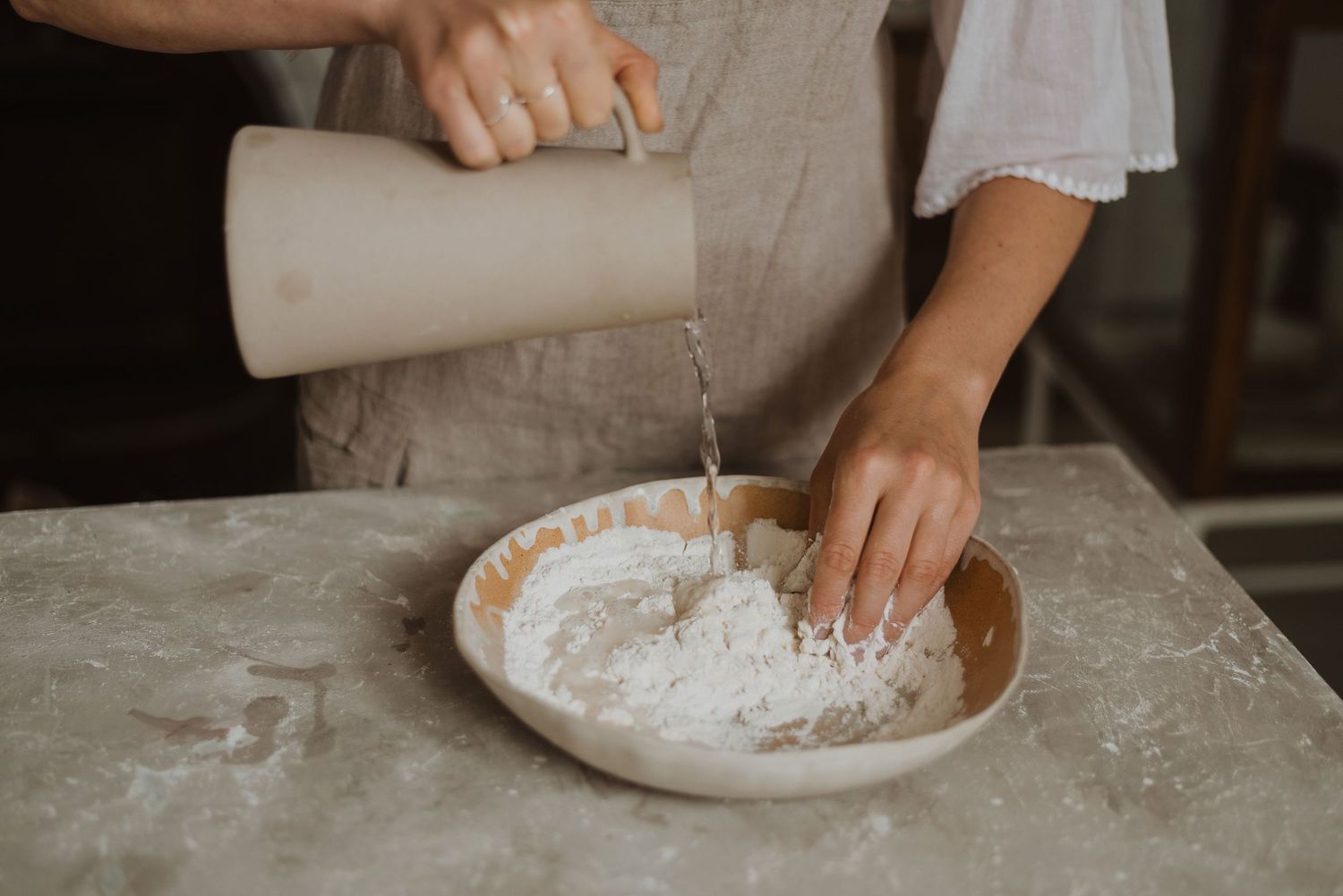 Person pouring water into flour in a ceramic bowl while baking bread on a marble countertop.