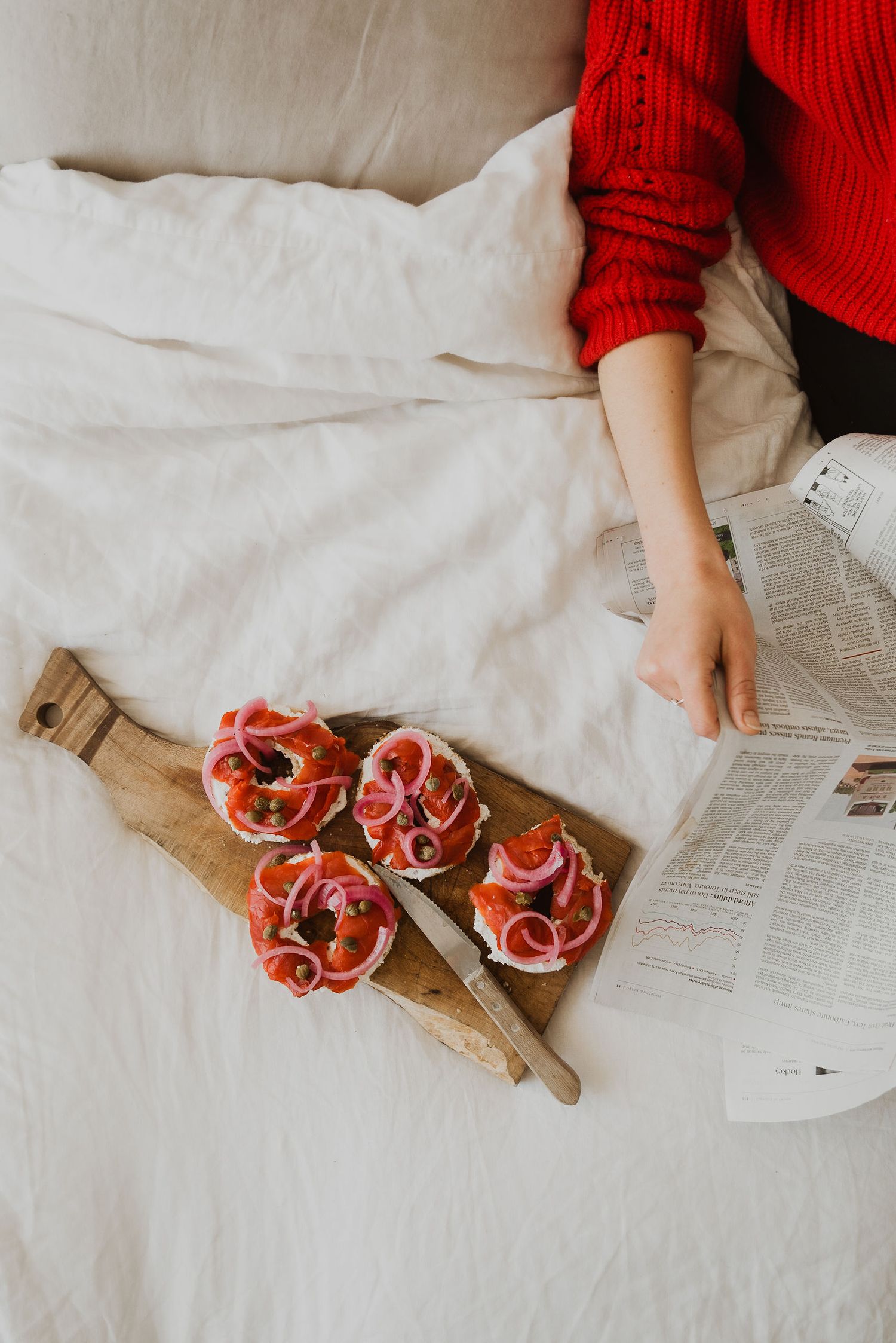 Person in red sweater reading newspaper in bed with smoked salmon open-faced sandwiches on a wooden cutting board.