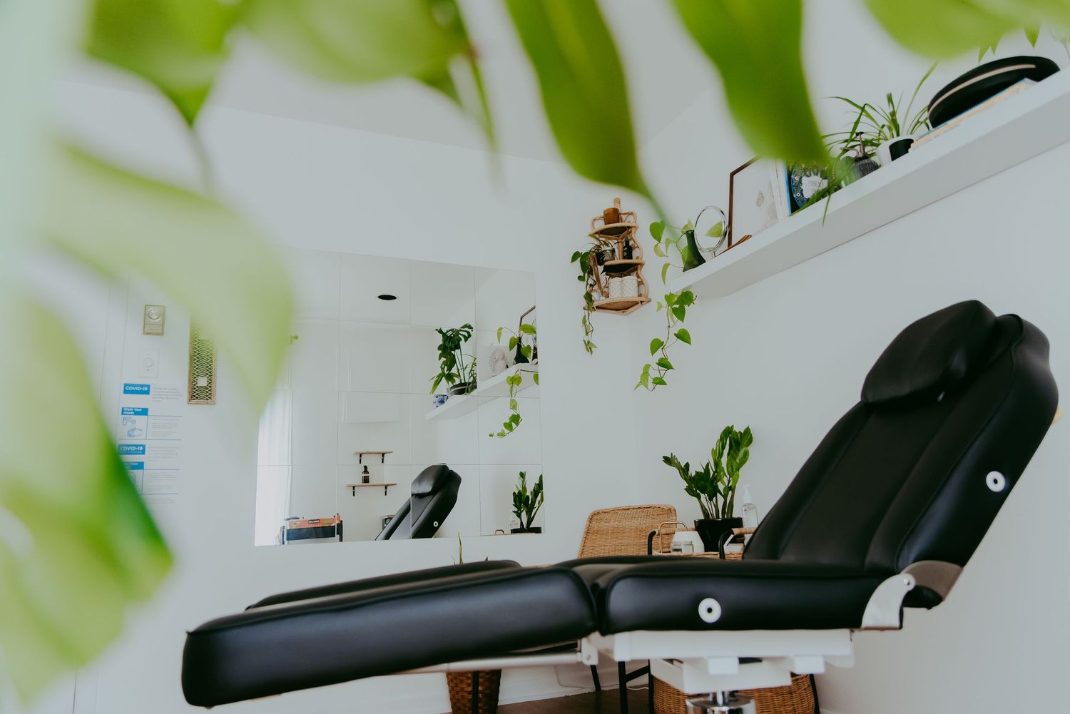 Modern massage therapy room with black treatment table, lush green plants, and white shelving in a bright, clean spa setting.