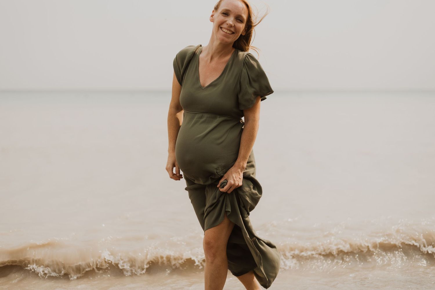 Smiling pregnant woman in olive green dress walking along sandy beach shore with gentle waves.