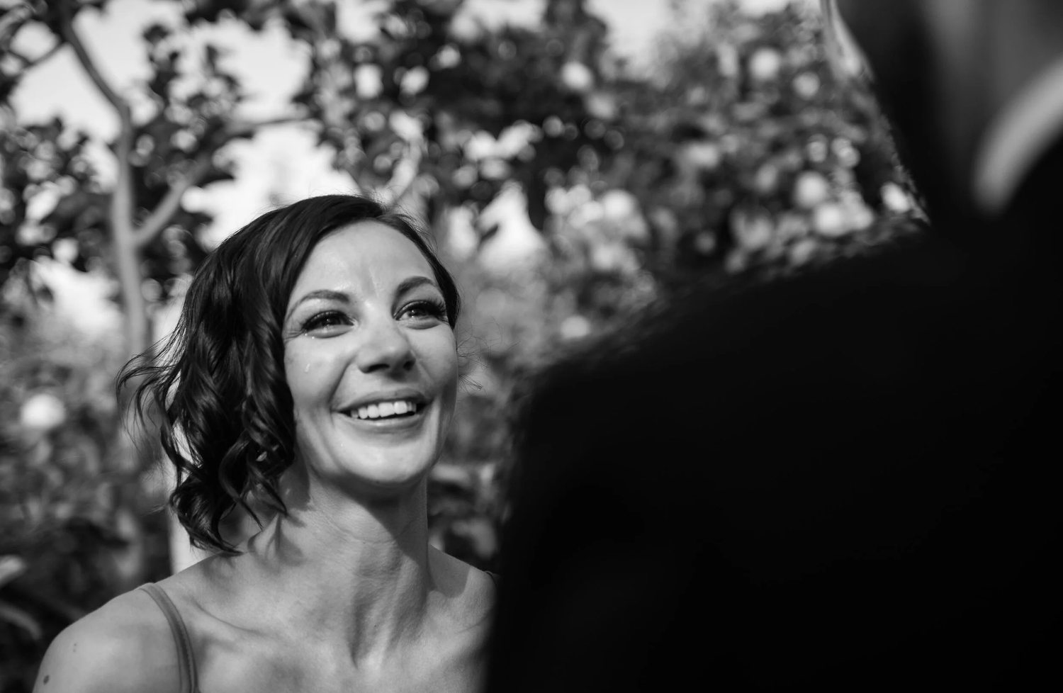 Bride smiling joyfully at groom during outdoor wedding ceremony in black and white photo.