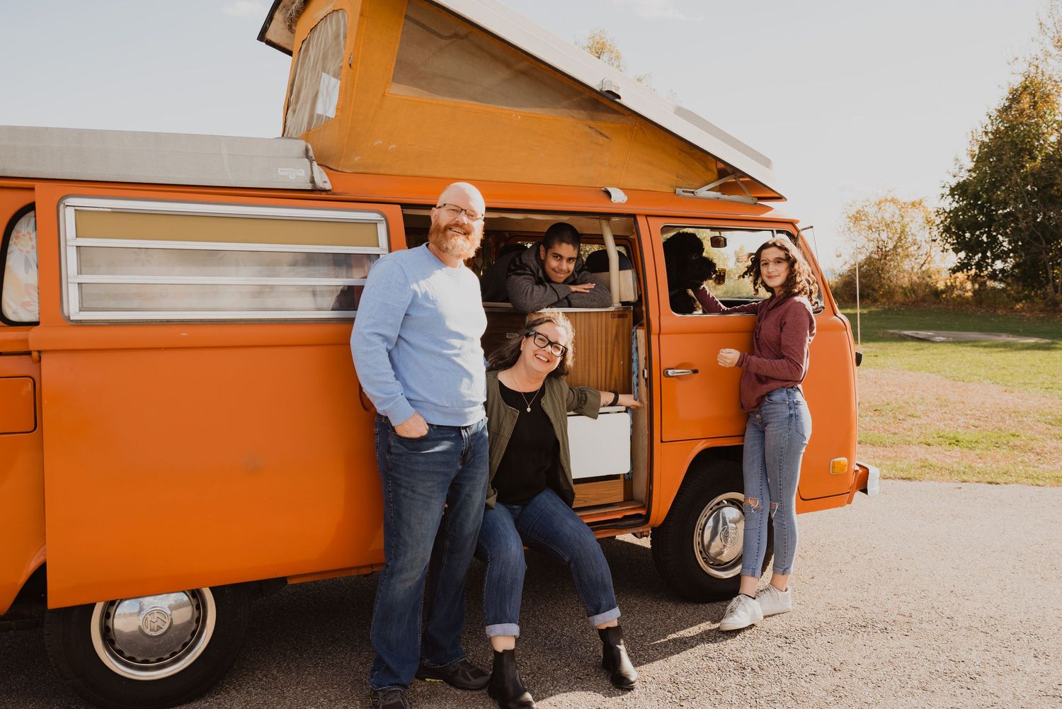 Four friends laughing beside a vintage orange VW camper van outdoors on a sunny day.