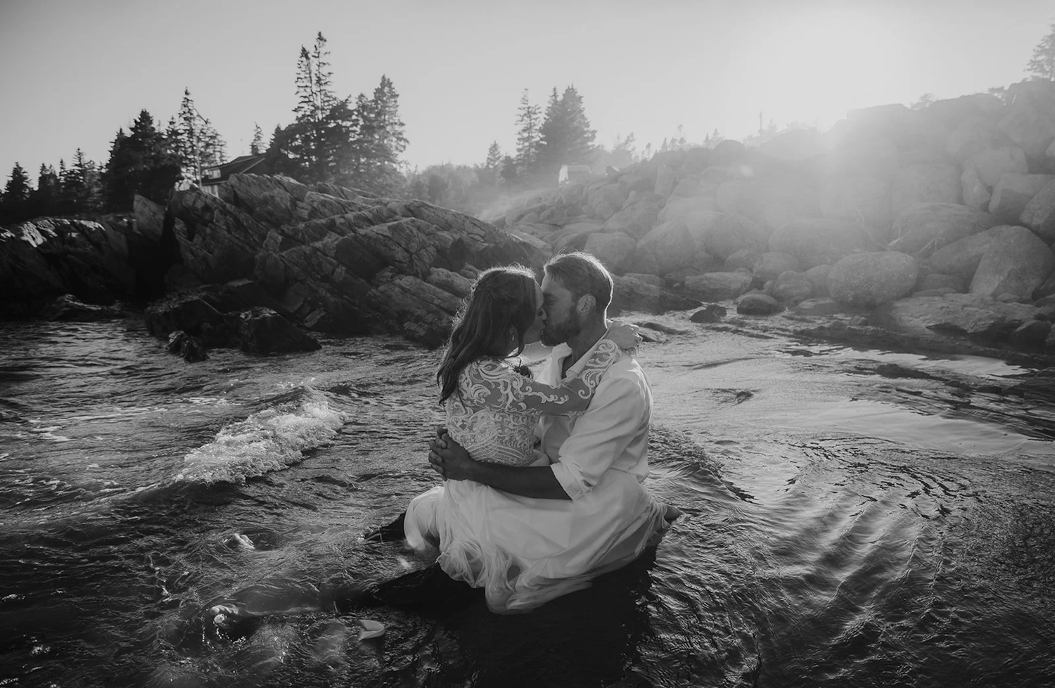 Black and white photo of a couple sharing a romantic kiss while sitting in a rocky stream with sunlight glowing behind them.