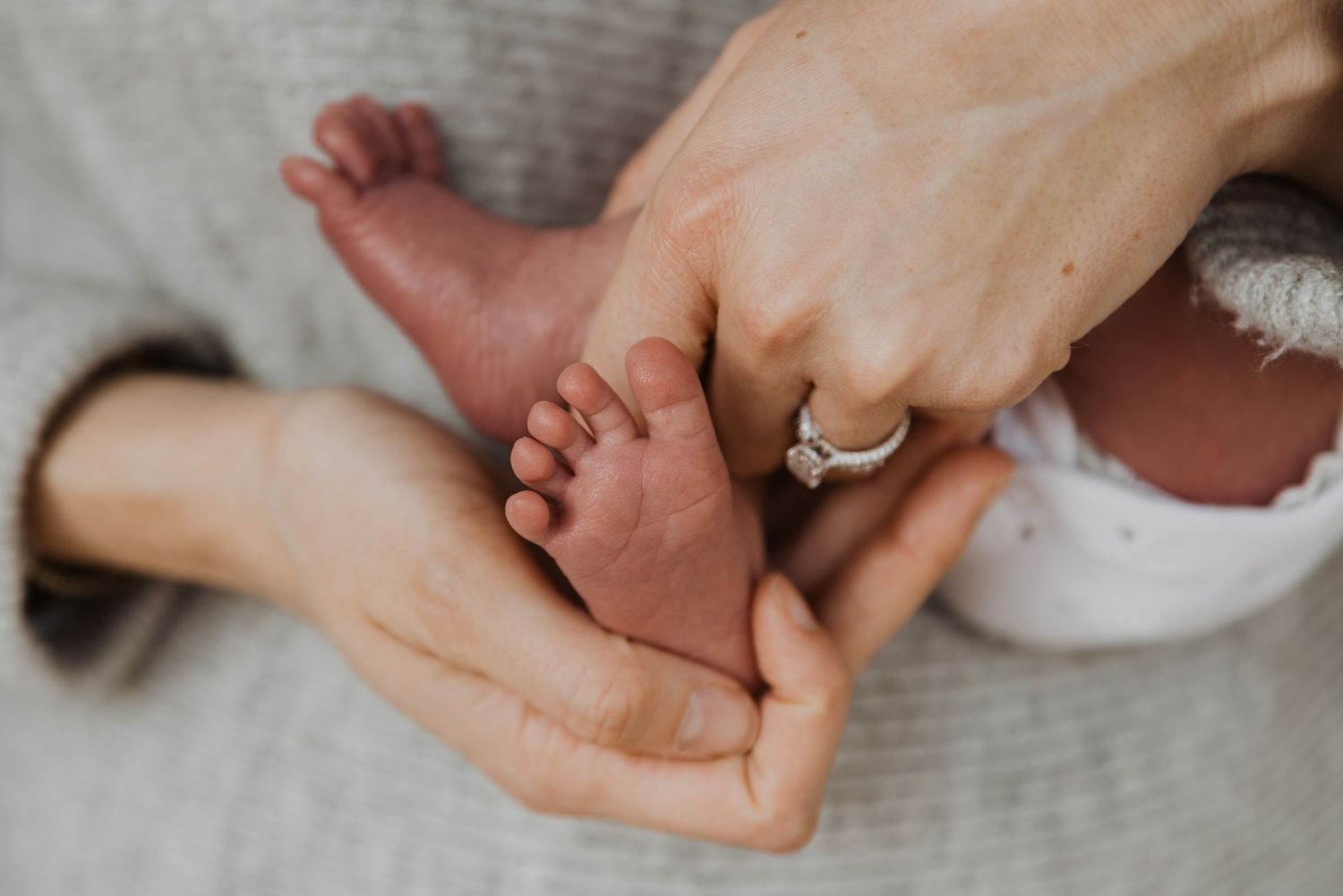 Close-up of newborn baby feet cradled in mother's hands, showcasing a diamond engagement ring.