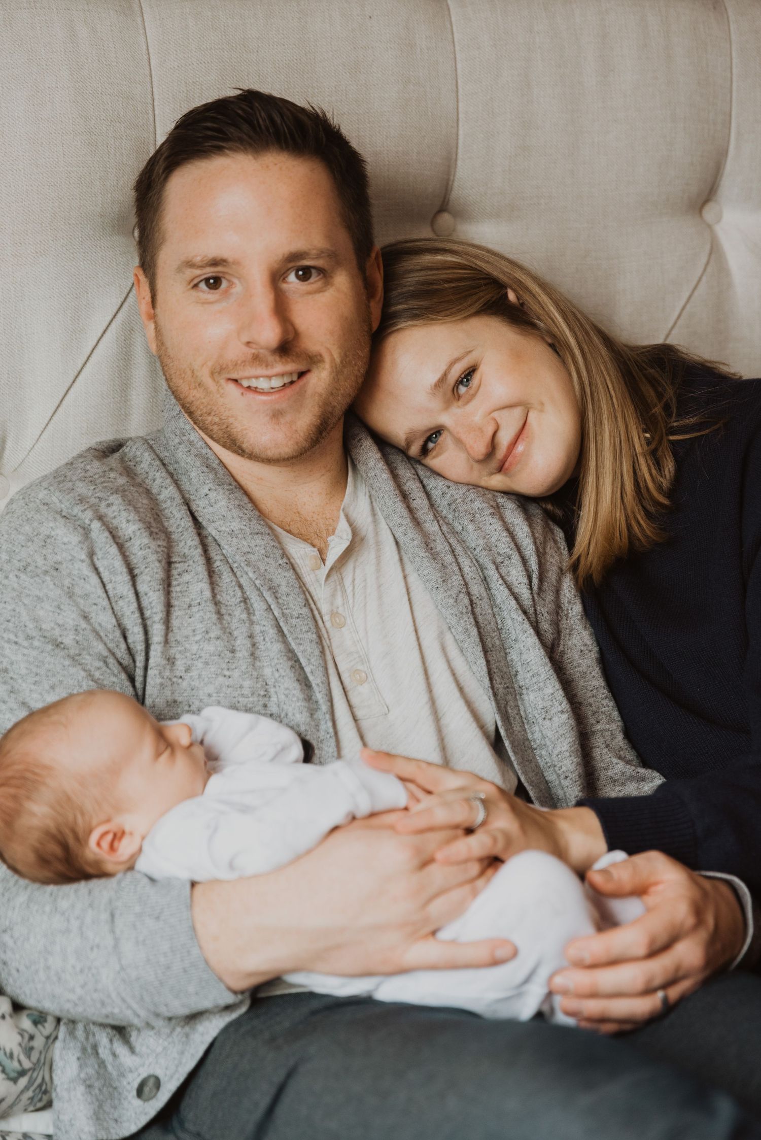 Smiling couple sitting on bed holding their sleeping newborn baby in a cozy home setting.