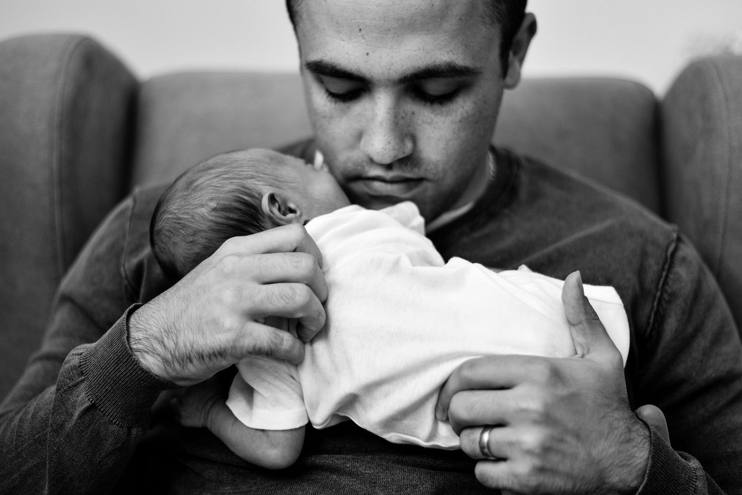 Black and white photo of a father tenderly cradling his newborn baby in his arms while seated on a sofa.