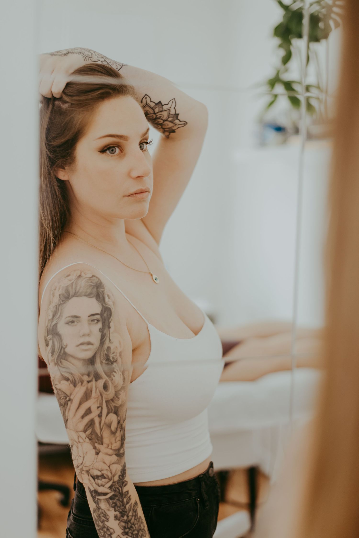 Young woman with sleeve tattoos poses in white crop top, hand raised to head, reflected in mirror.
