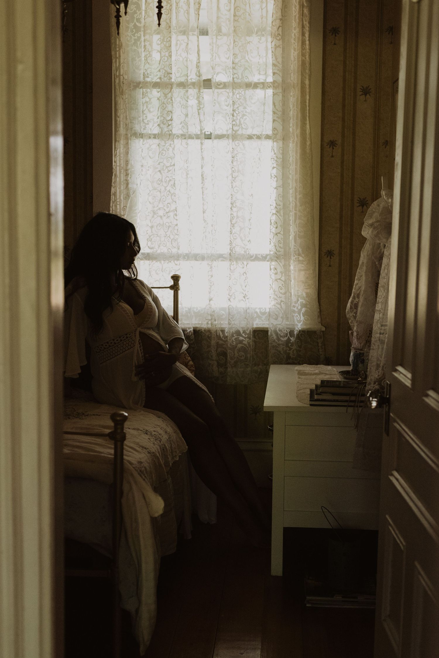 A woman in white lingerie sits on a bed by a lace-curtained window in a warmly lit vintage bedroom.