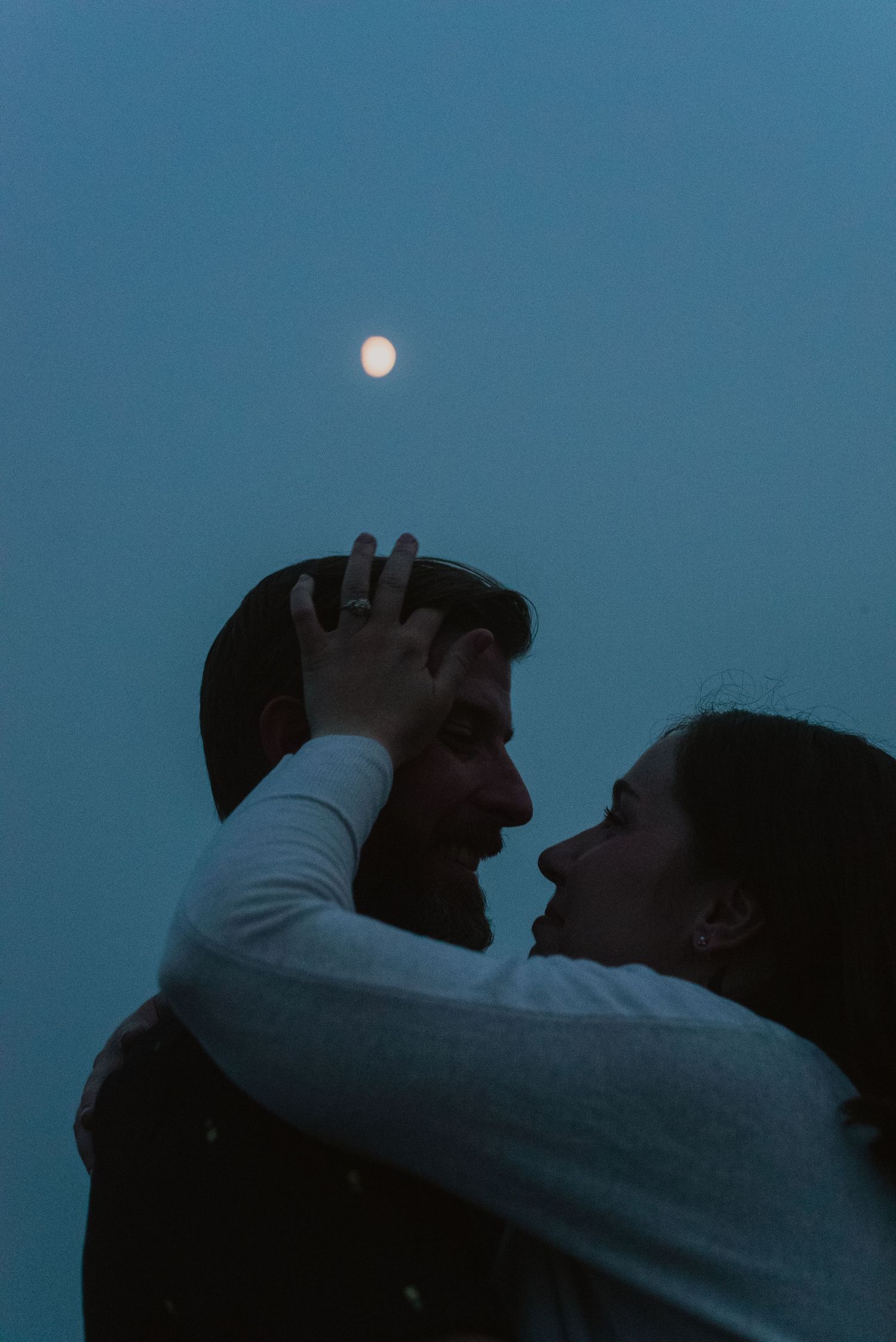 Romantic couple embracing under full moon at dusk, woman holding mans face tenderly against blue sky.