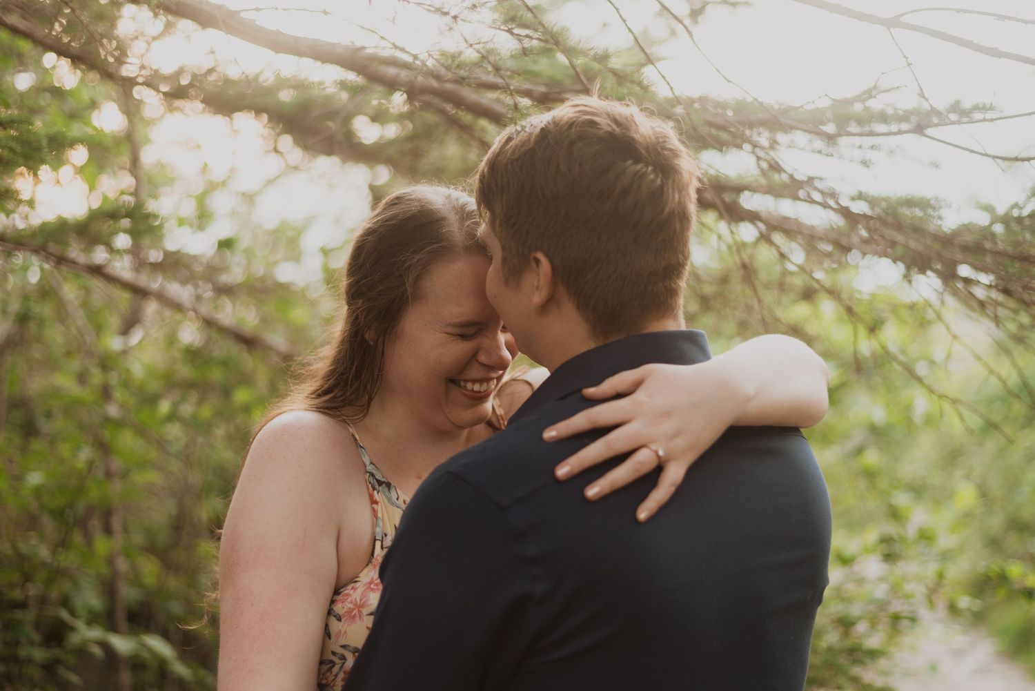 A joyful couple embraces outdoors, laughing together under lush green trees in a romantic engagement photo.